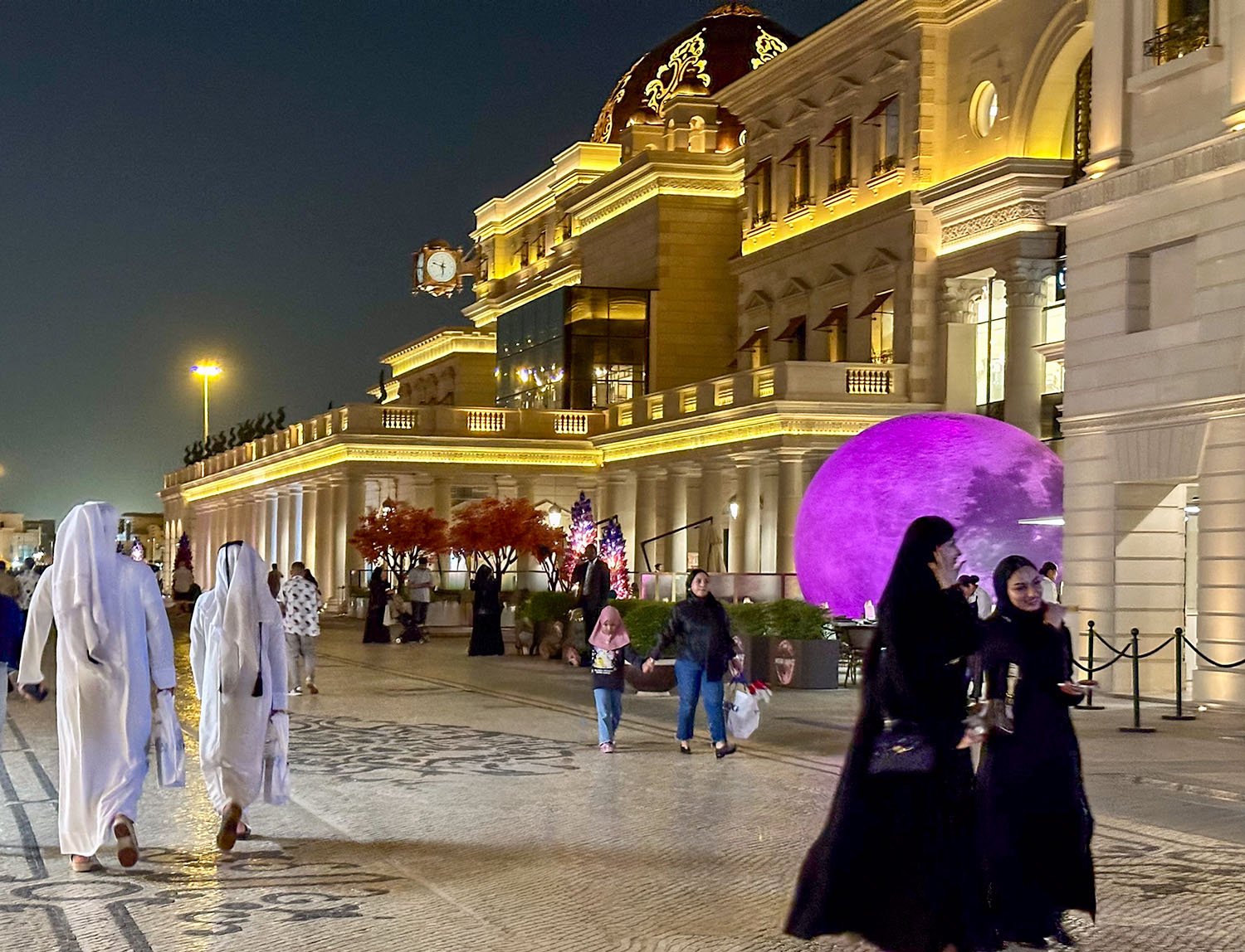 Qatari citizens stroll along a wide boulevard between ornate buildings at nighttime in Doha, Qatar..
