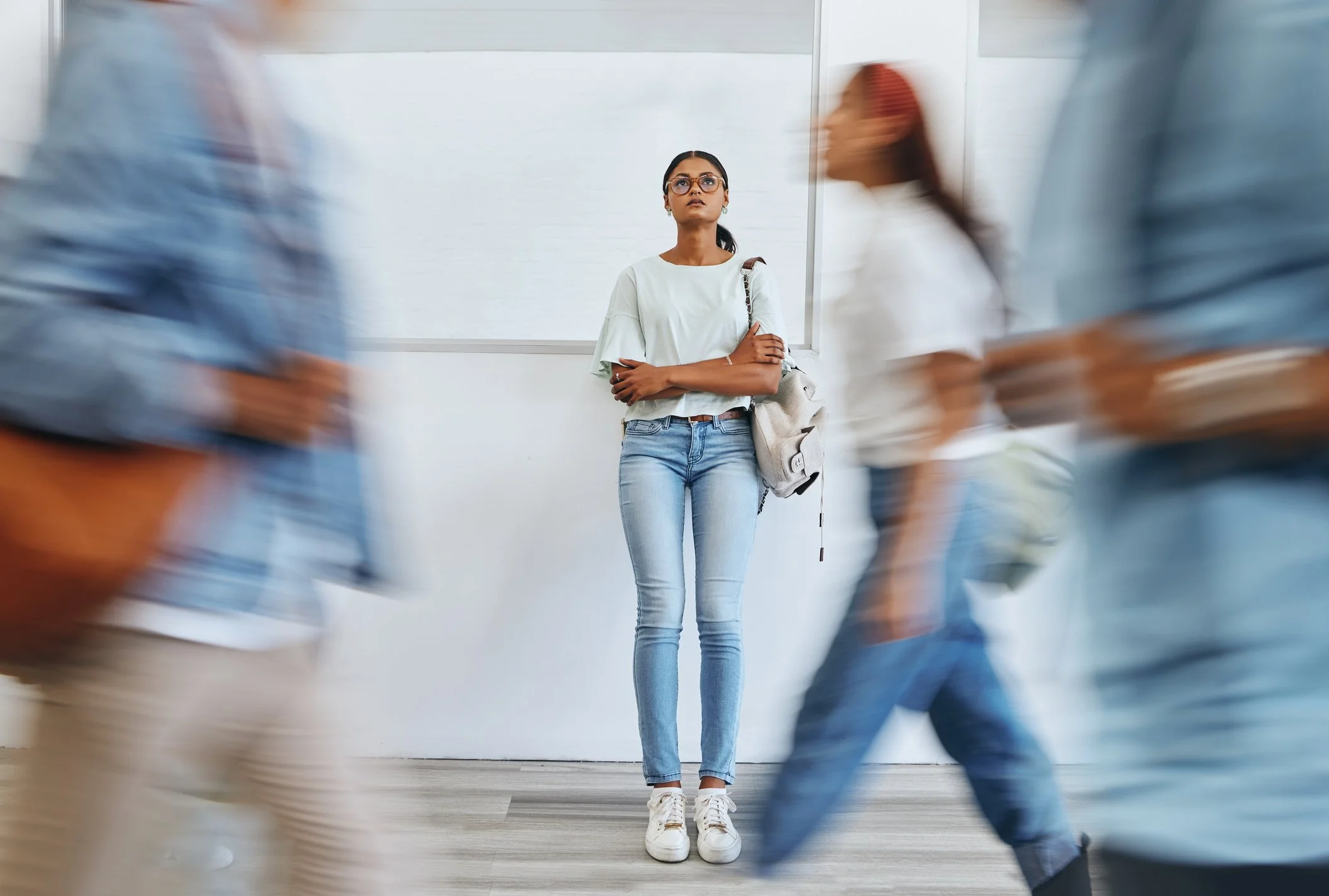 Young woman with glasses and a backpack standing with arms crossed as people pass by in motion blur in a hallway.
