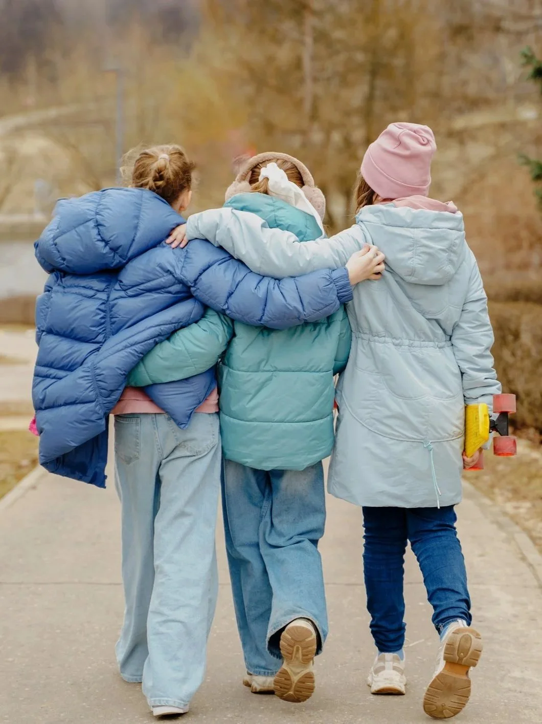 Three friends with their arms around each other walking on a path in autumn, wearing puffer jackets and jeans, with one holding a skateboard.