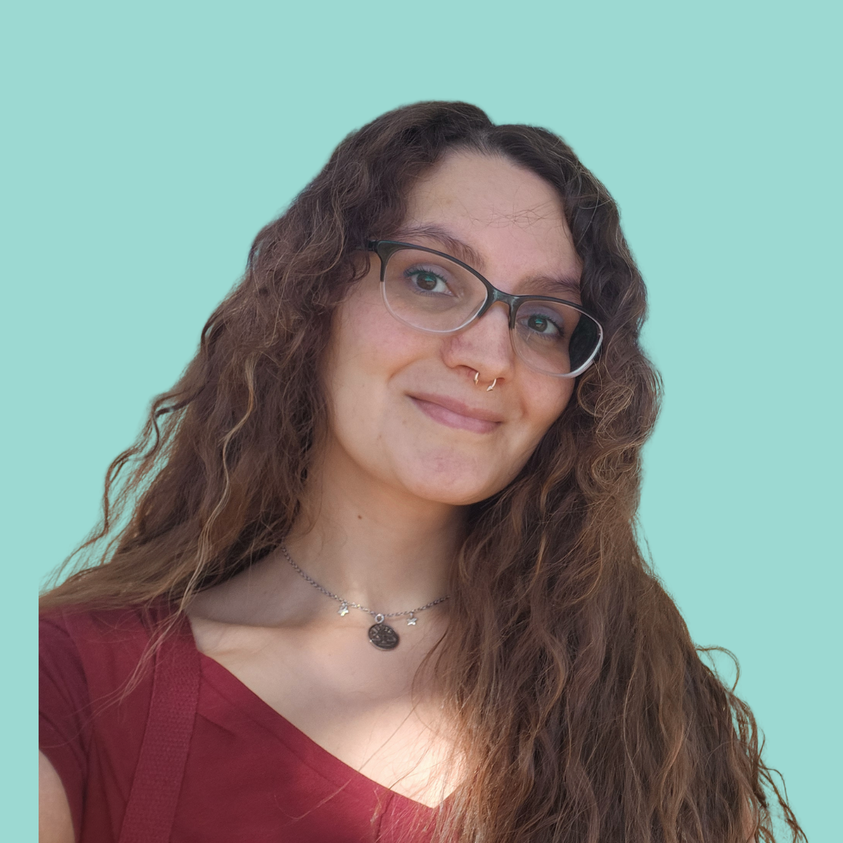 A woman with long curly brown hair with purple streaks, wearing glasses, a pink beanie, a matching pink scarf, and a zip-up jacket, smiling at the camera against a light blue background.