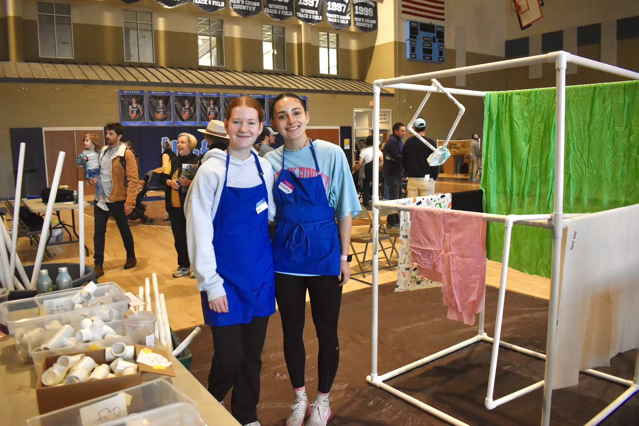 Two volunteers in aprons next to a PVC pipe house at Playhouse BuildFest.
