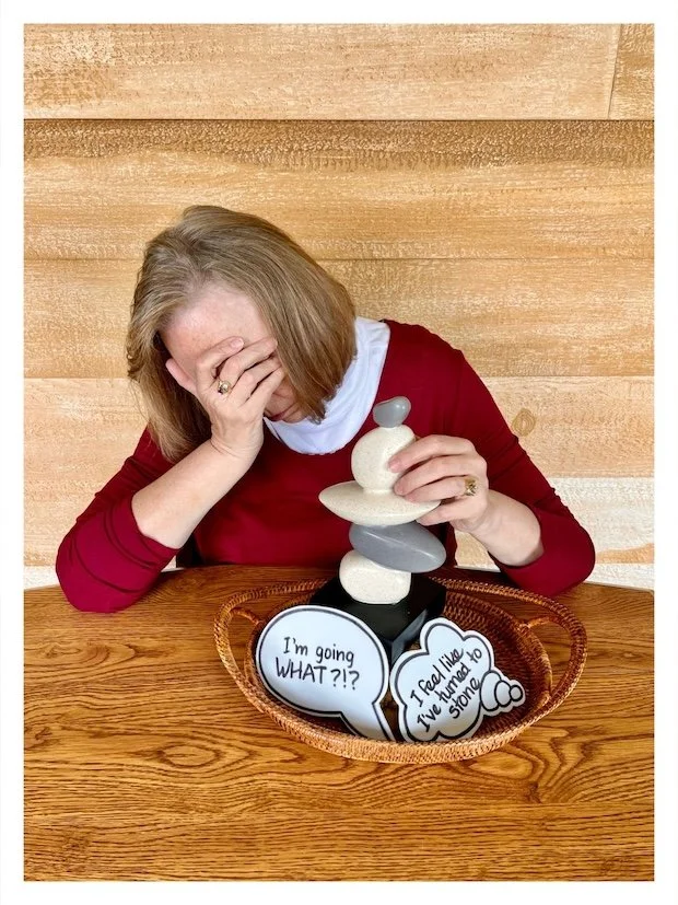 Centered on a basket is a stacked rock cairn. Two cutout cartoon bubbles are leaning against the rocks. One bubble says: "I'm going WHAT?!?" The other says: "I feel like I've turned to stone." Linda's posture represents exhaustion and dispair.