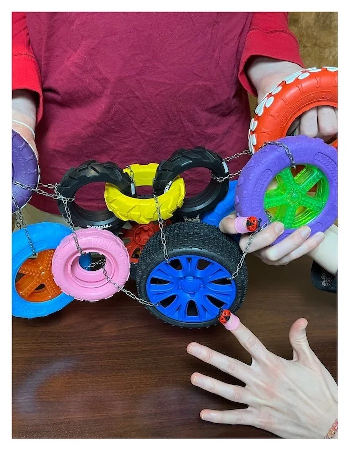 A pair of hands are wearing ladybug finger puppets. Above is a wild colorful bundle of toy tires, chained together. They're descending on the ladybugs, nearly crushing them. One hand is trying to grab a tire, the other appears startled.