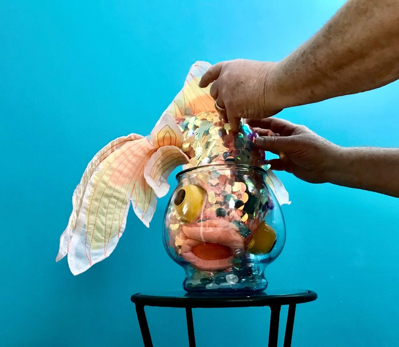 A big silly goldfish is stuck in a fishbowl that's too small for it. It's shoved headfirst into the bowl with its fins extending out the top. It has bulging eyes and a crooked mouth expressing discontent. Two hands are feeling the fish.