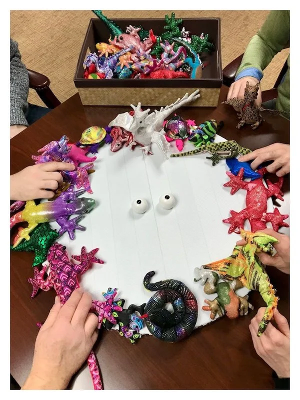A flat white circular base, representing a visual field, fills most of the photo. A pair of eyeballs are in the center, Six hands are reaching in from all sides. The hands are placing the little whimsical toy monsters around the periphery.