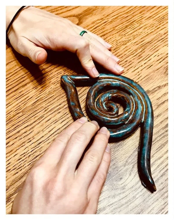 A pair of hands are feeling a stylized eye made from a coil of clay. A central spiral creates the pupil, then wraps under for the lower lid. A point shapes the tear duct. The coil sweeps above and beyond the pupil, creating the mascara line.