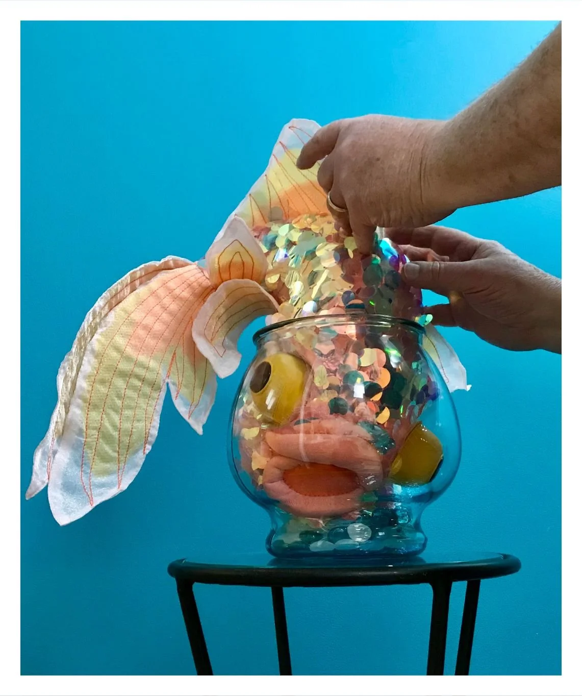 A big silly toy goldfish is stuck in a fishbowl that's too small for it. It's shoved headfirst into the bowl with its tail and fins extending out the top. It has bulging eyes and a crooked mouth expressing discontent. Two hands are feeling the fish.