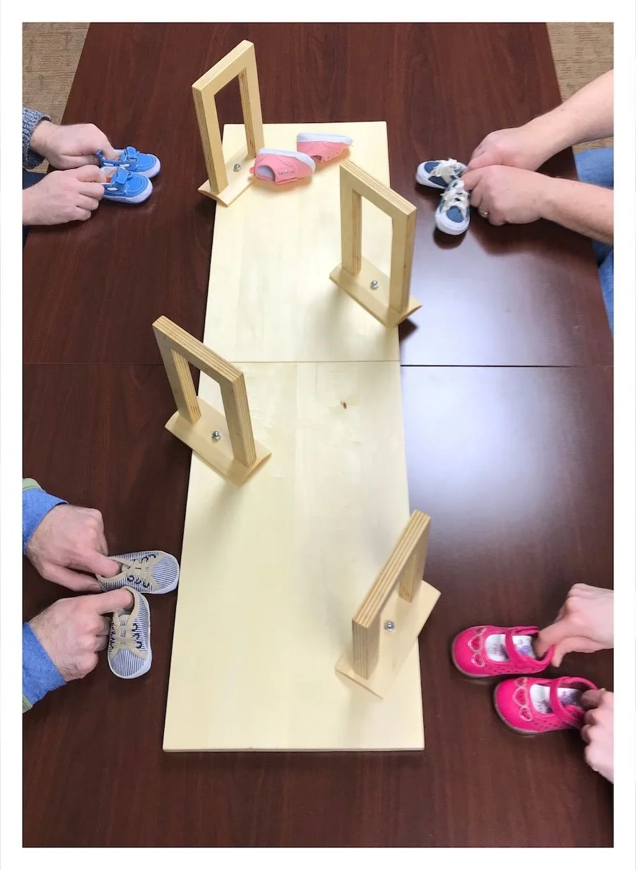 A wooden model of a hall with four doorways along the sides, runs through the photo. The walls and rooms are left to the imagination. The doorways are cocked at goofy angles. Eight hands are interacting with the model. They're holding baby shoes.