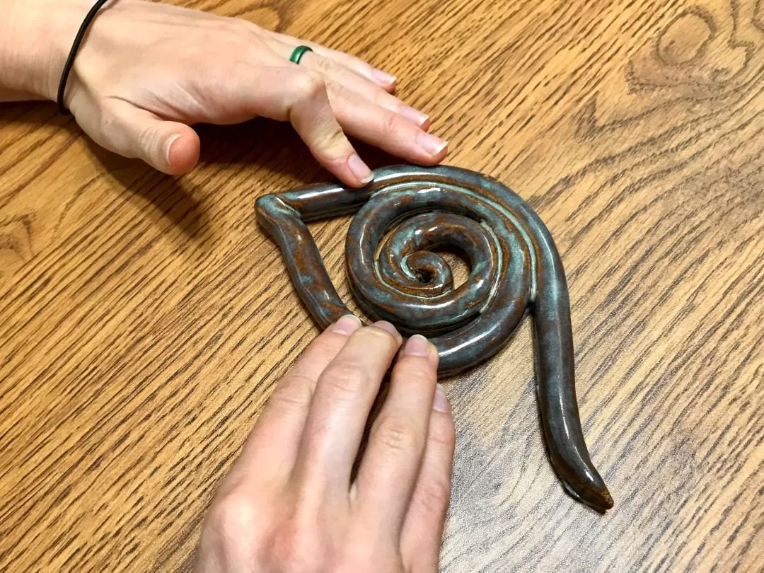 A pair of hands are feeling a stylized eye made from a coil of clay. A central spiral creates the pupil, then wraps under for the lower lid. A point shapes the tear duct. The coil sweeps above and beyond the pupil, creating the mascara line.