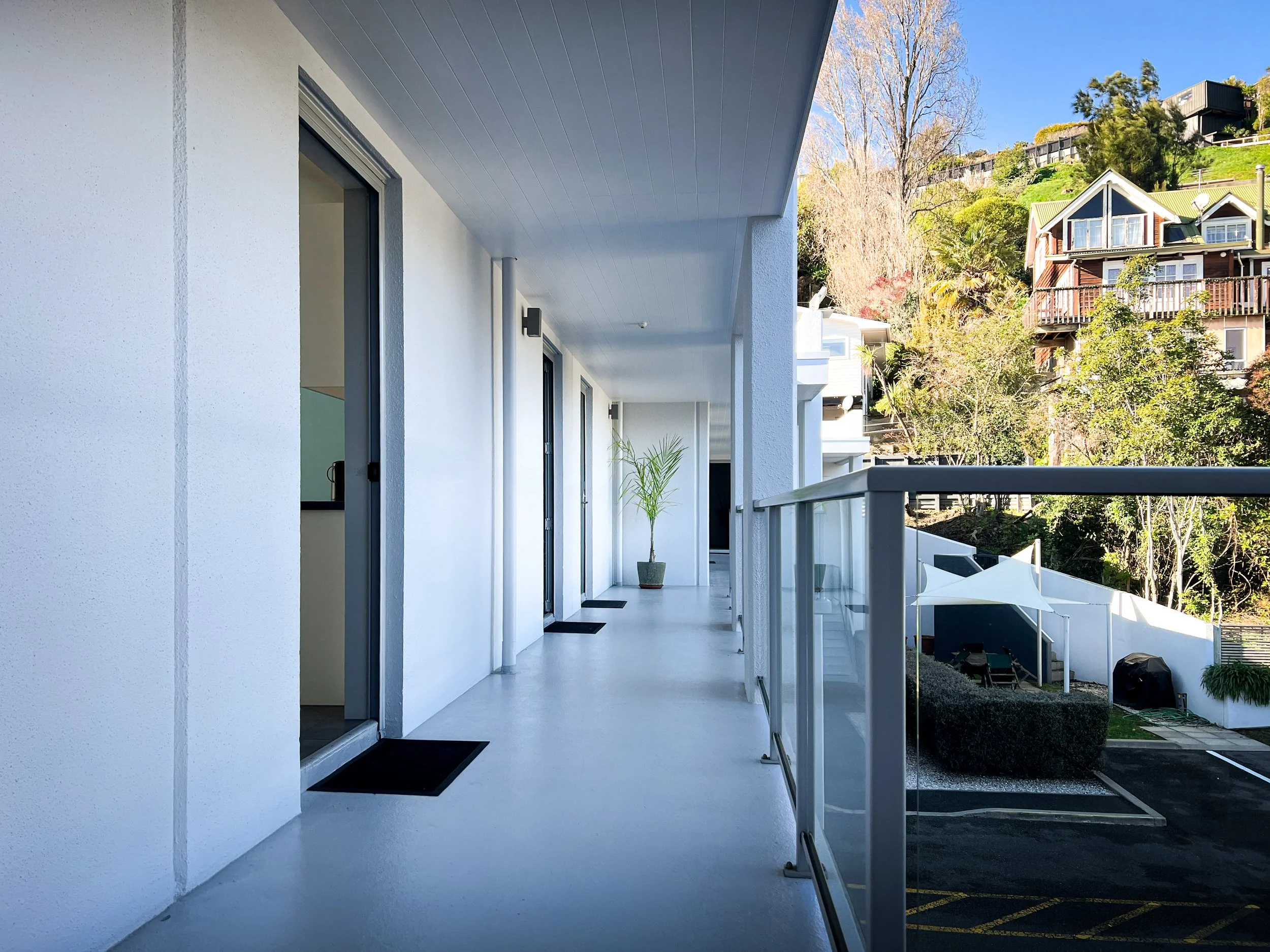 Apartment balcony with white walls, sliding glass doors, and black mats, overlooking a parking lot and hilly residential area with houses surrounded by trees.