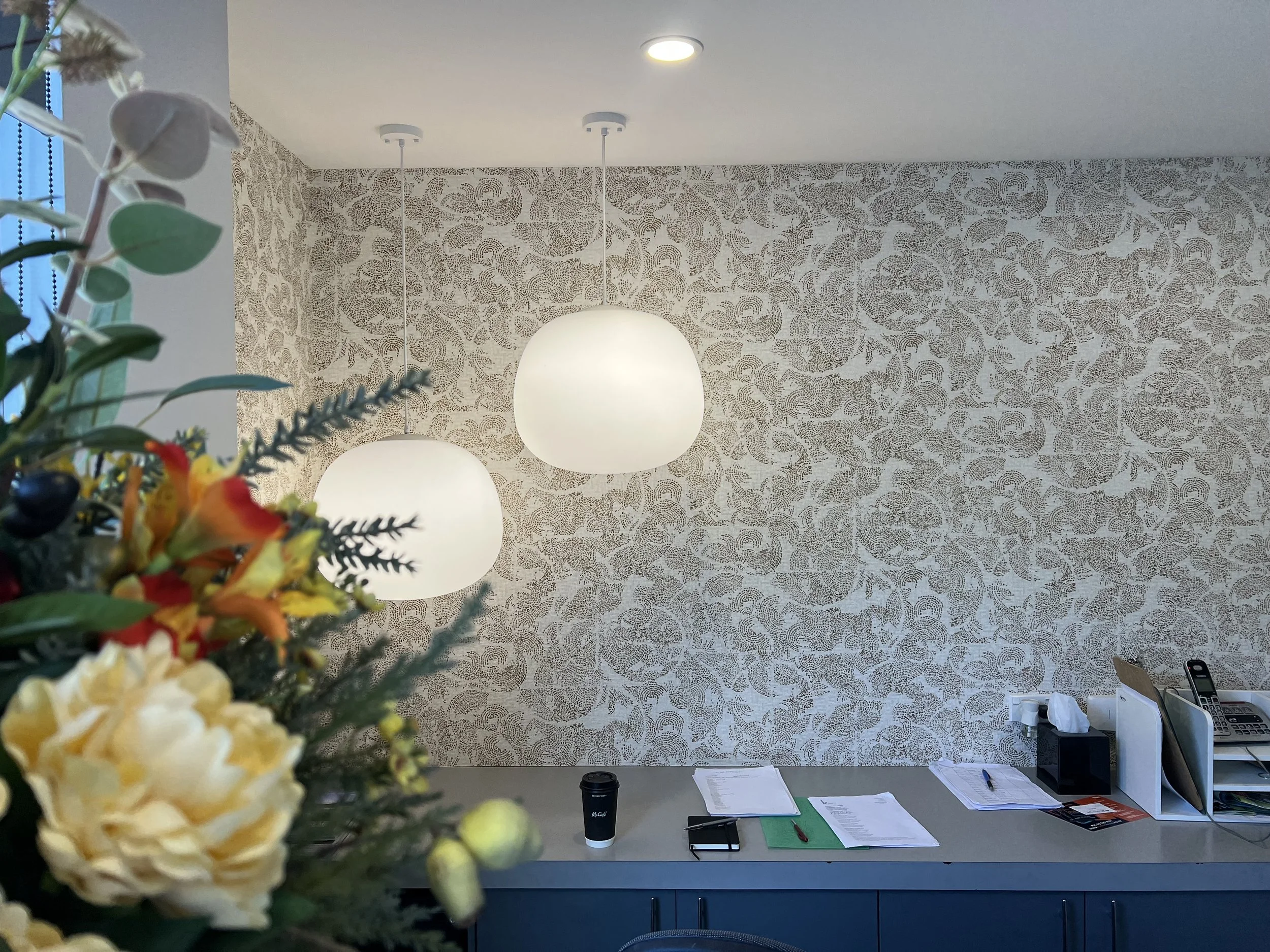 Office reception area with a floral arrangement in the foreground, a desk with papers, pens, a coffee cup, and office supplies against a patterned wallpapered wall with two hanging white globe lights.