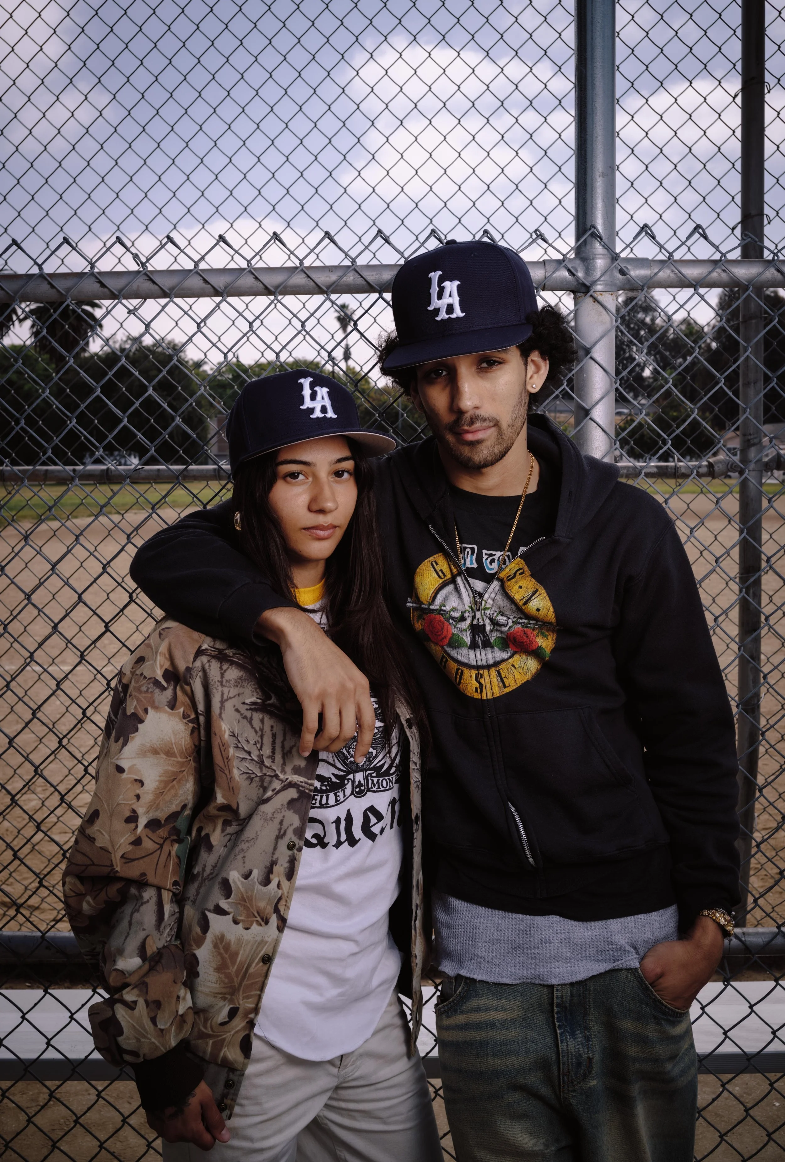 A young woman and a young man are standing in front of a chain-link fence at a baseball field, both wearing Los Angeles Dodgers caps, with trees and a cloudy sky in the background.