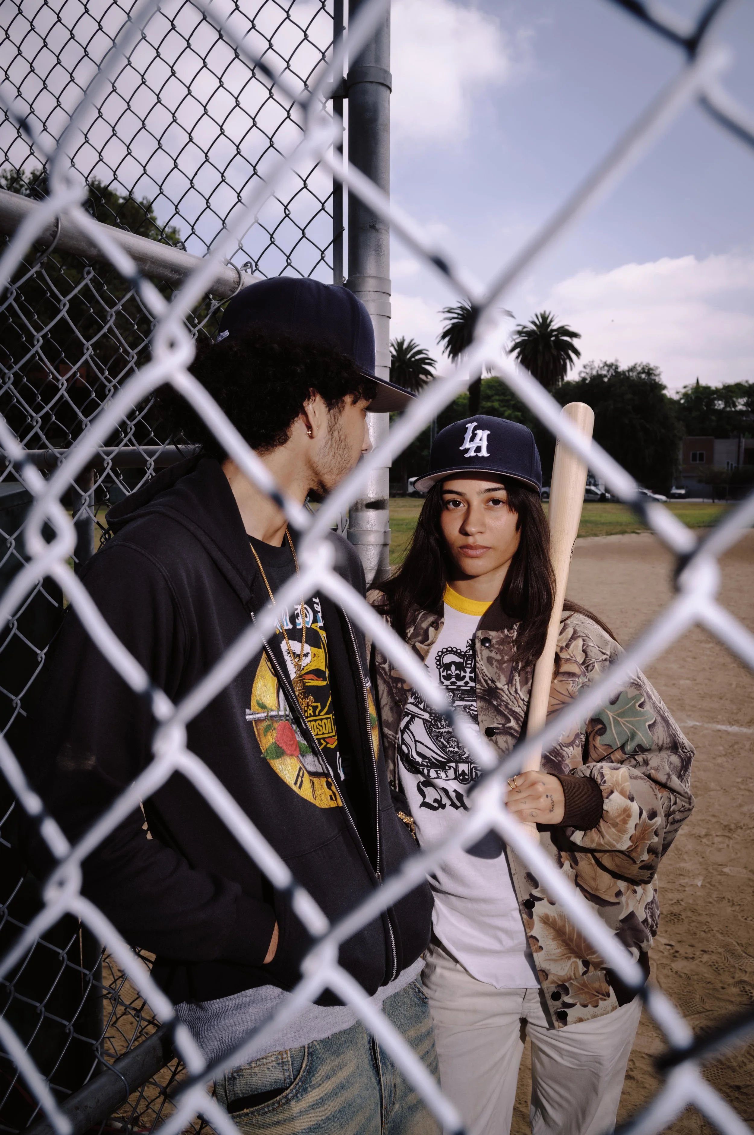 Two young women stand behind a chain-link fence at a baseball field, with palm trees and a partly cloudy sky in the background. One woman holds a baseball bat and wears a Los Angeles Dodgers cap, while the other is dressed in camouflage clothing and a white T-shirt.