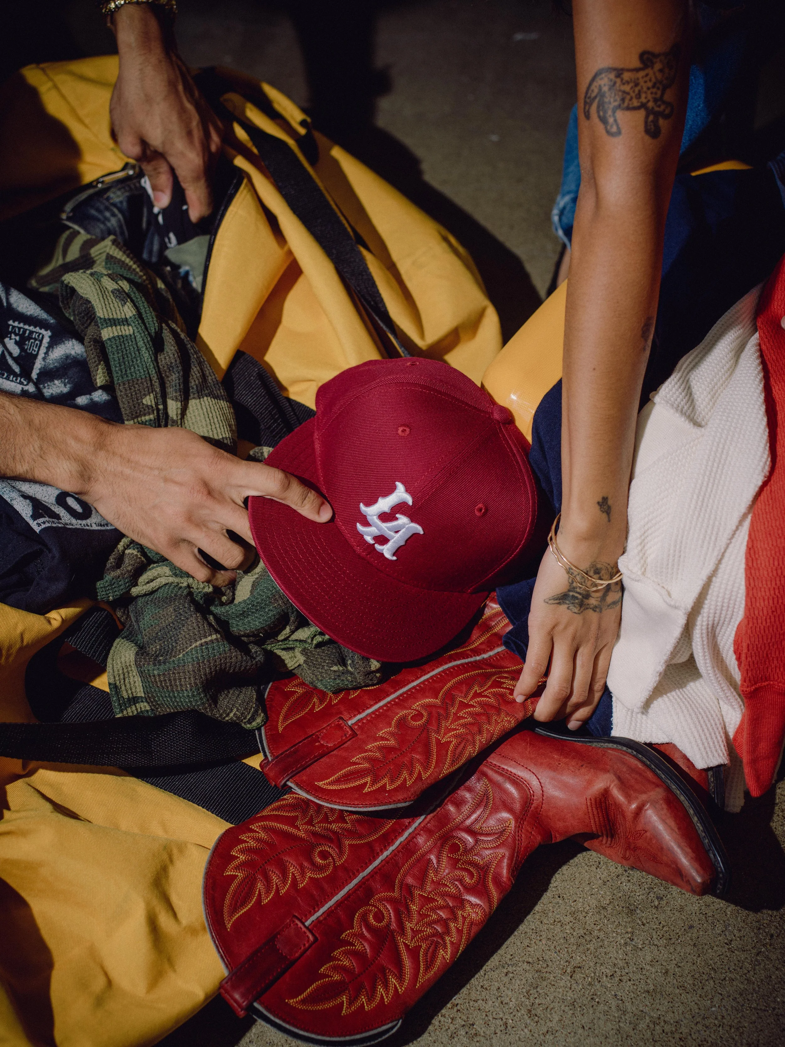 People sitting on the ground, with various bags and clothing, including a red baseball cap with a white logo and a tattooed arm.