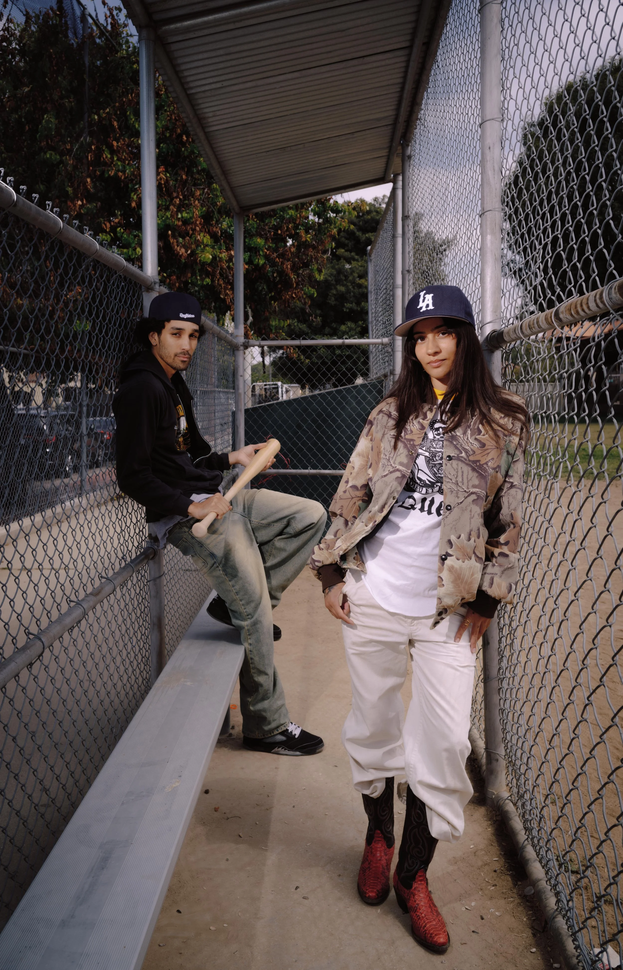 Two young adults, a man and a woman, are at a baseball field, with the woman standing in front and the man sitting on a bench in the background. They are both wearing baseball caps.