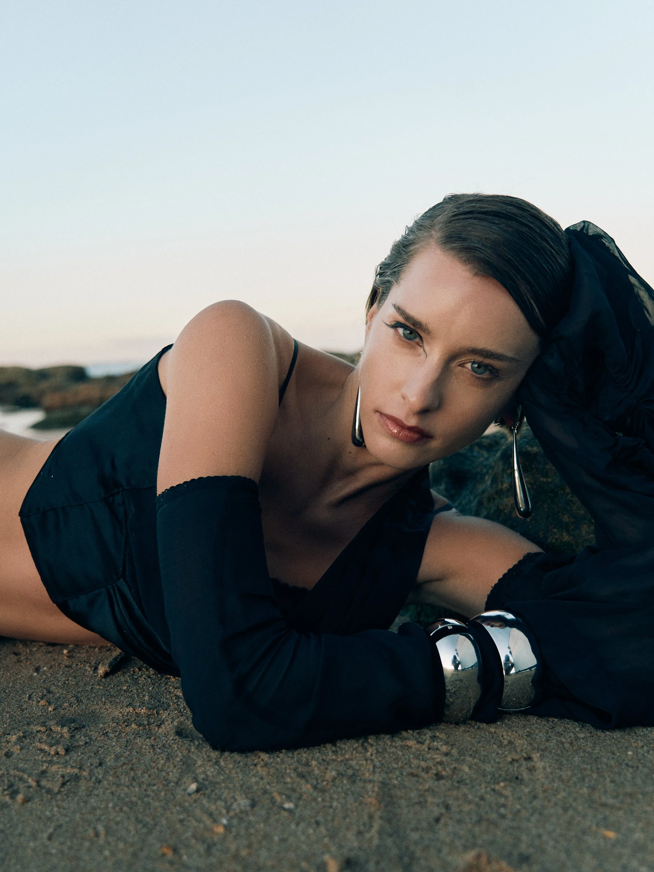 A woman with dark hair lying on a sandy surface outdoors, dressed in black, with one arm propped up and the other arm bent near her head, looking directly at the camera with a serious expression.
