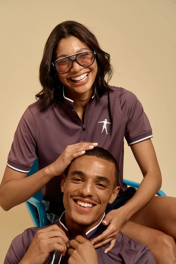 Two smiling young adults, a woman and a man, posing together. The woman has medium-length dark hair, glasses, and is wearing a purple athletic shirt. She is sitting on the man's shoulders, touching his head with one hand. The man has short dark hair,