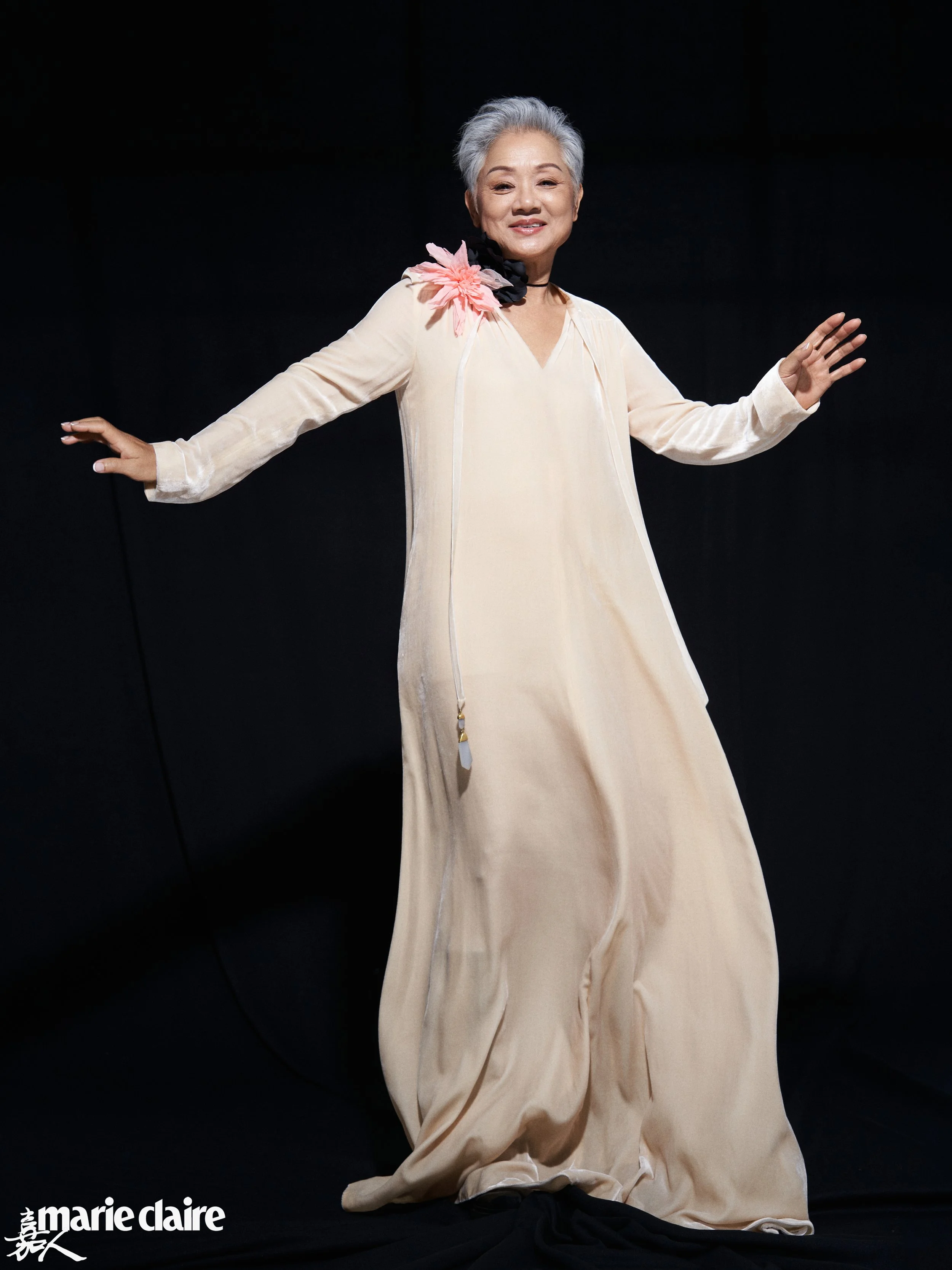 An elderly woman with gray hair wearing a long, beige dress with a pink flower on her shoulder, smiling with her arms outstretched against a black background.