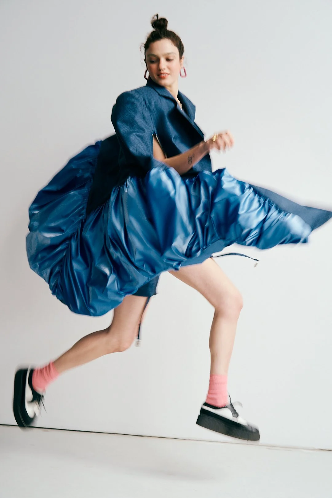 A young woman in a blue jacket and shiny blue skirt jumping against a plain white background.
