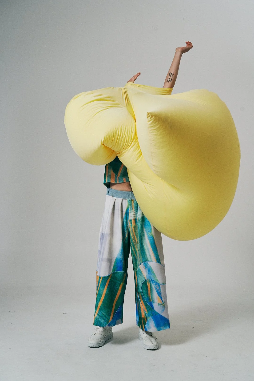 Person holding large yellow inflatable object obscuring their body, standing against a plain light gray background, wearing colorful wide-leg pants and white shoes.