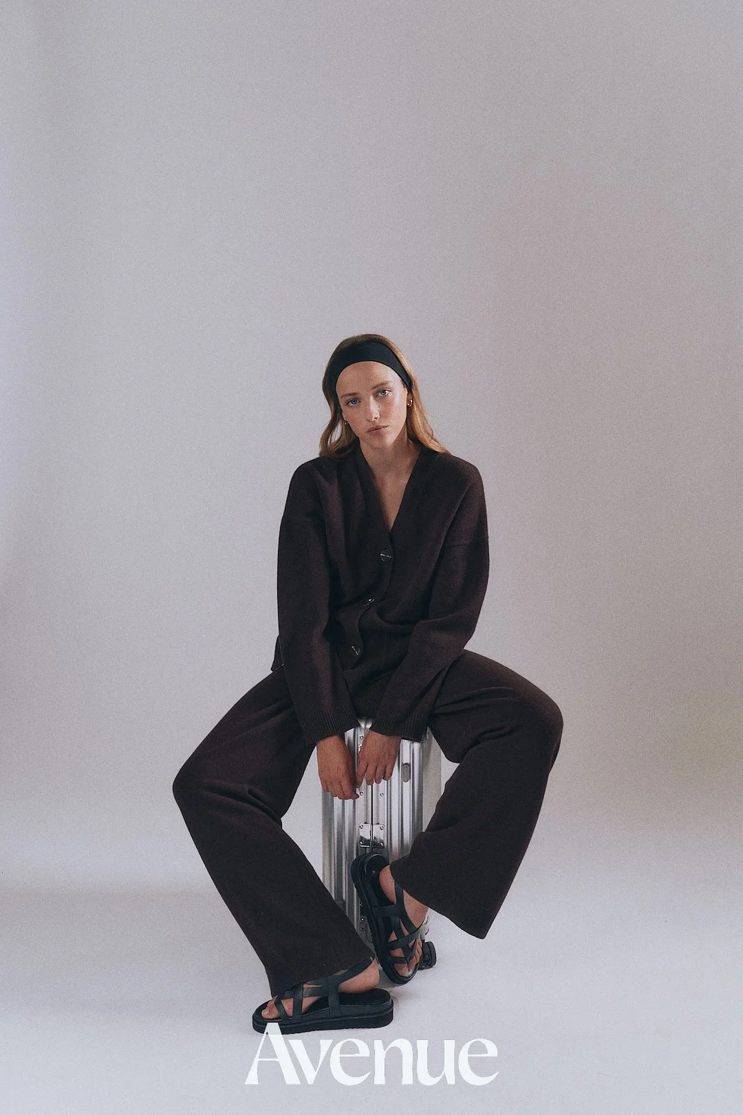 A woman with light skin and long wavy hair, wearing a black headband, black oversized cardigan, loose black pants, and black sandals, sitting on a silver suitcase against a plain light gray background, with the word 'Avenue' at the bottom.