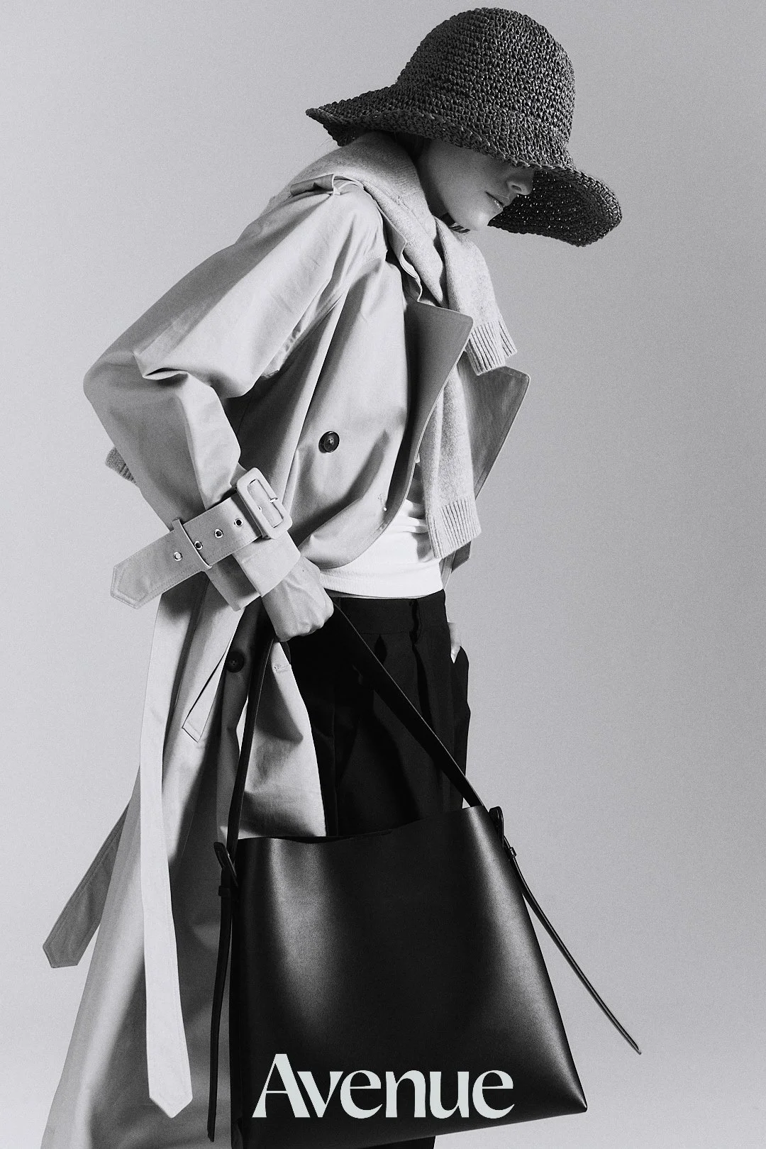 A black and white photo of a woman wearing a wide-brimmed hat, trench coat, scarf, and black pants, carrying a large black shopping bag with the word 'Avenue' printed on it.