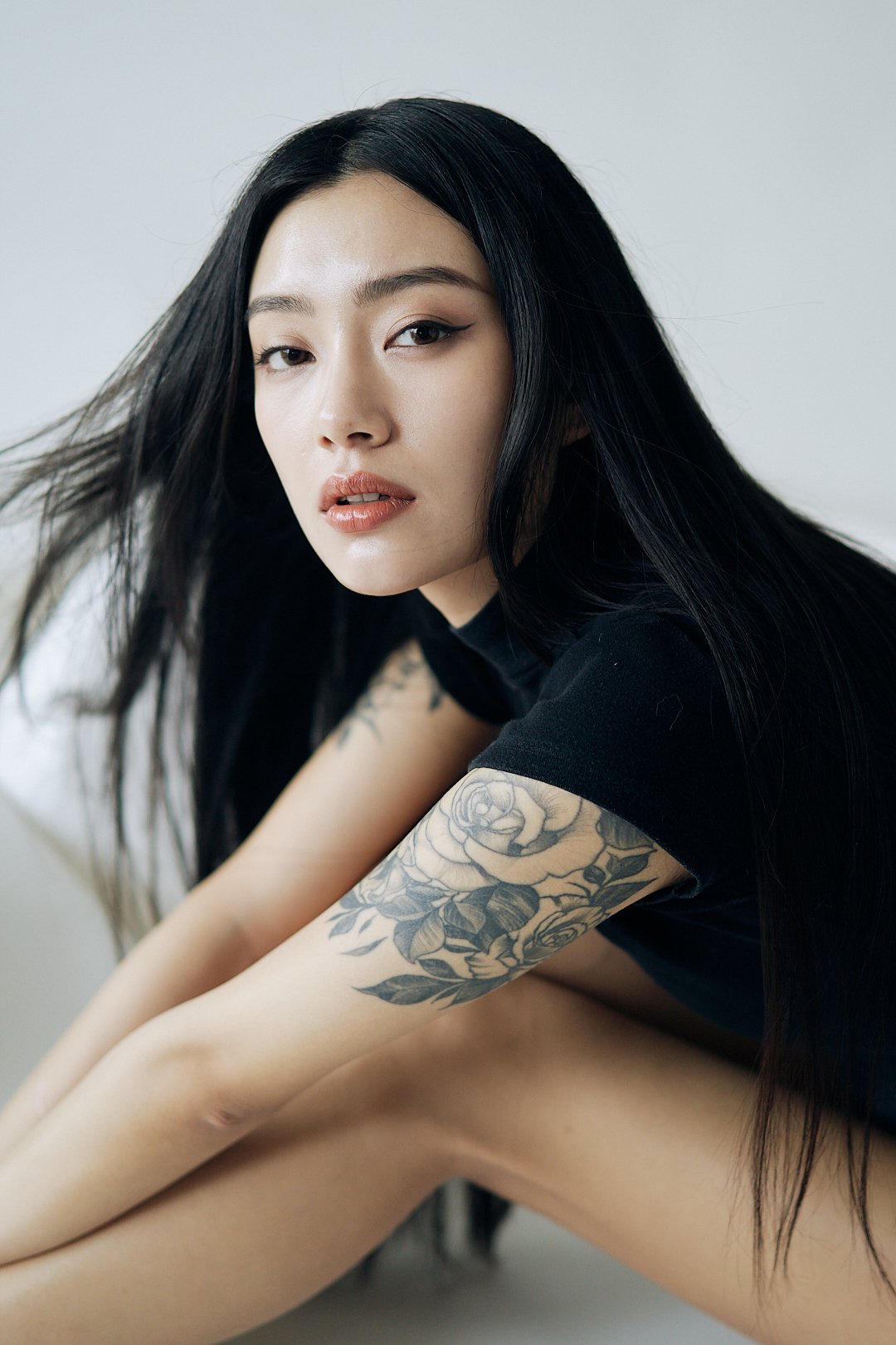 A woman with long black hair, wearing a black top, sitting against a neutral background. She has a tattoo of roses on her left arm and is looking at the camera.