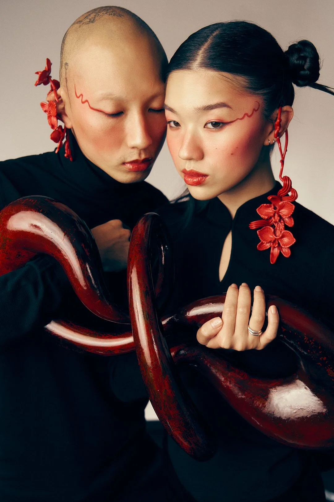 Two women with Asian features dressed in black, holding a large, glossy red and black octopus sculpture, with red floral earrings and makeup with red lines.