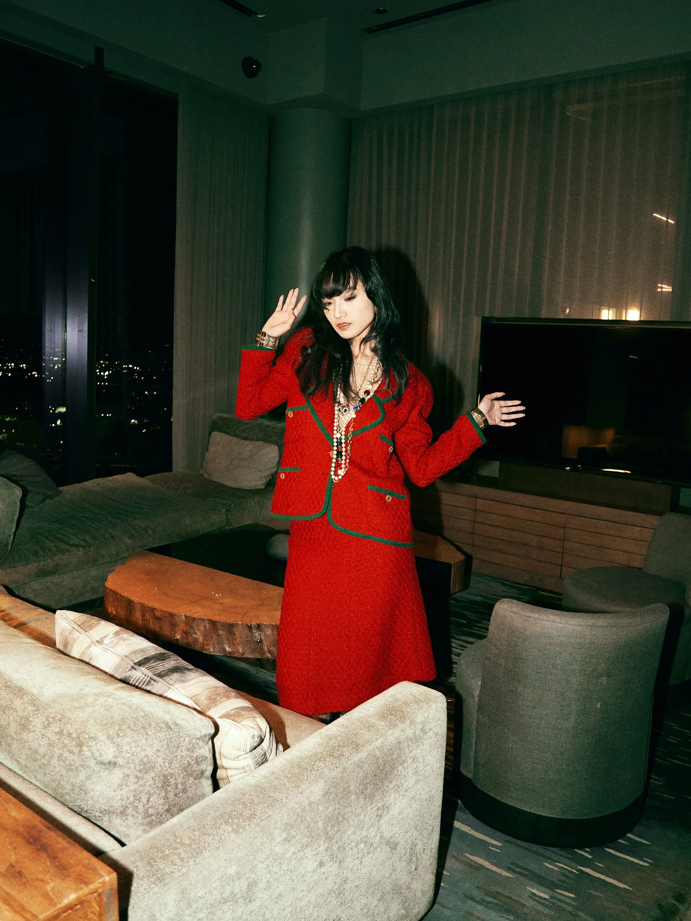 Woman in a red outfit with jewelry standing in a modern living room at night.