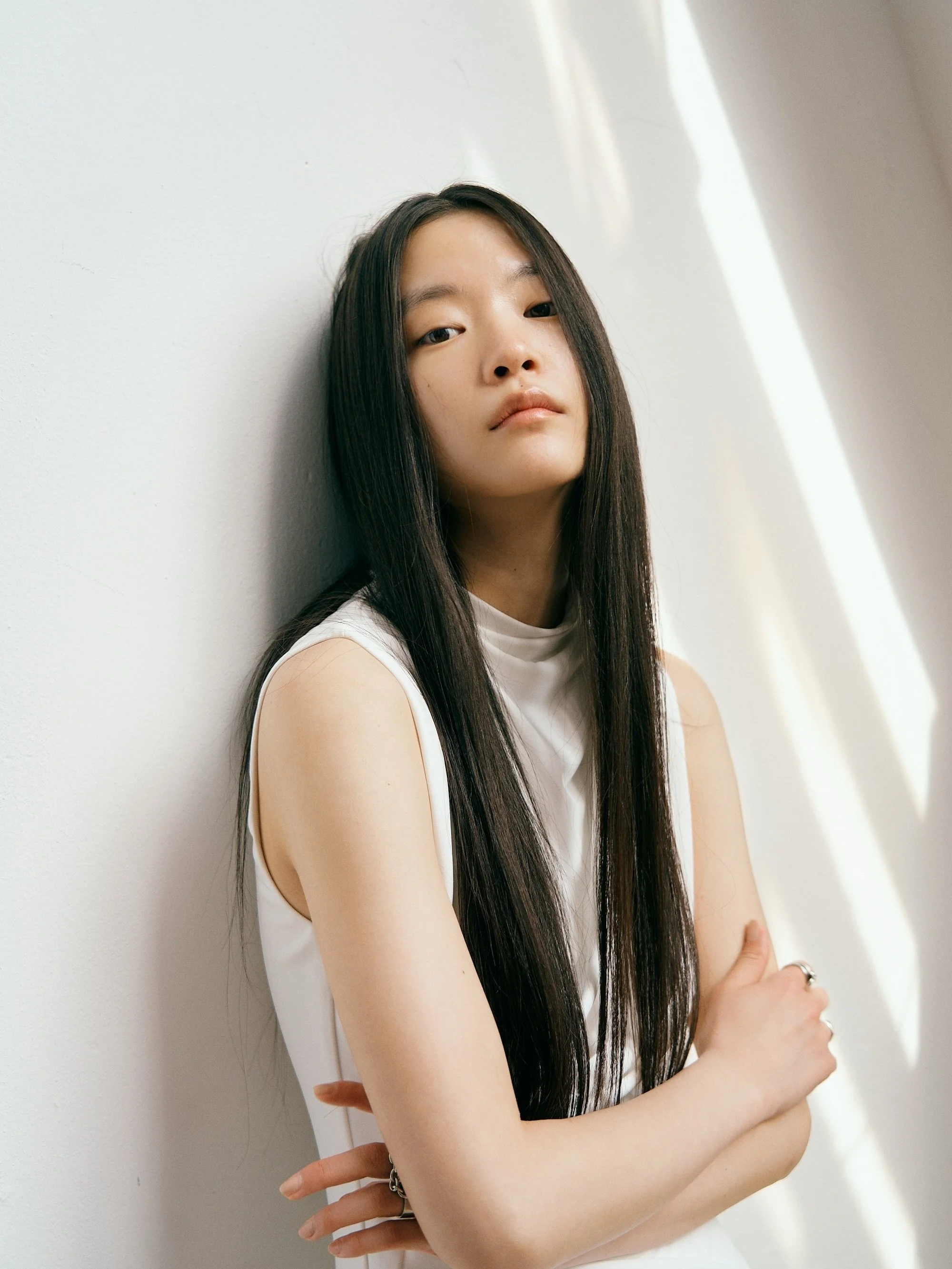 A young woman with long dark hair leaning against a painted white wall, wearing a sleeveless white top, with sunlight creating shadows on the wall.