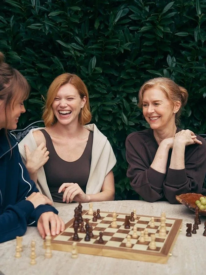 Three women sitting at a table playing chess outdoors, laughing and enjoying each other's company against a backdrop of green foliage.