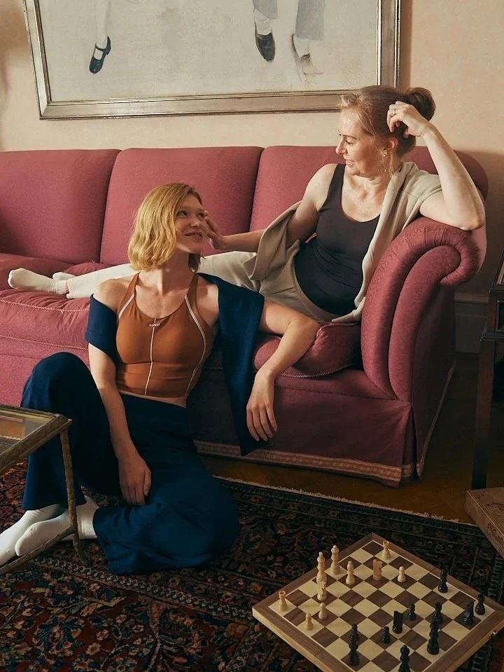 Two women in a cozy living room, one sitting on the floor with a chessboard in front of her, and the other reclining on a pink sofa, smiling and touching the other woman’s face.