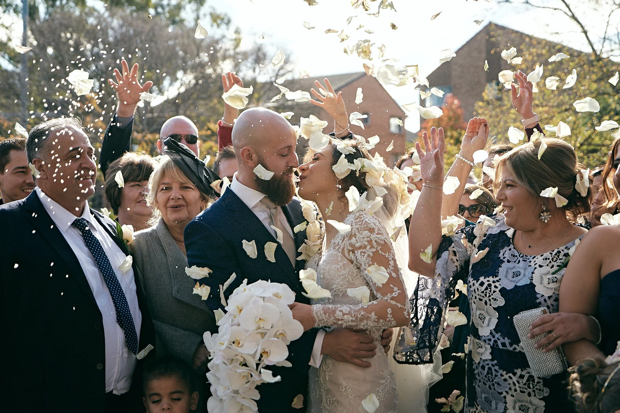 Wedding couple kissing surrounded by friends and family, celebrating with flower petals in the air outdoors during daytime.