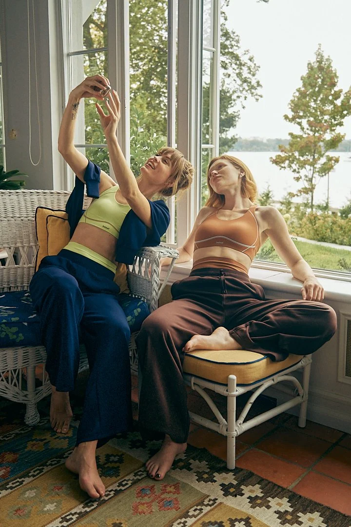 Two young women sitting by a large window, taking a selfie together, with a view of trees and water outside, wearing casual, comfortable clothing.