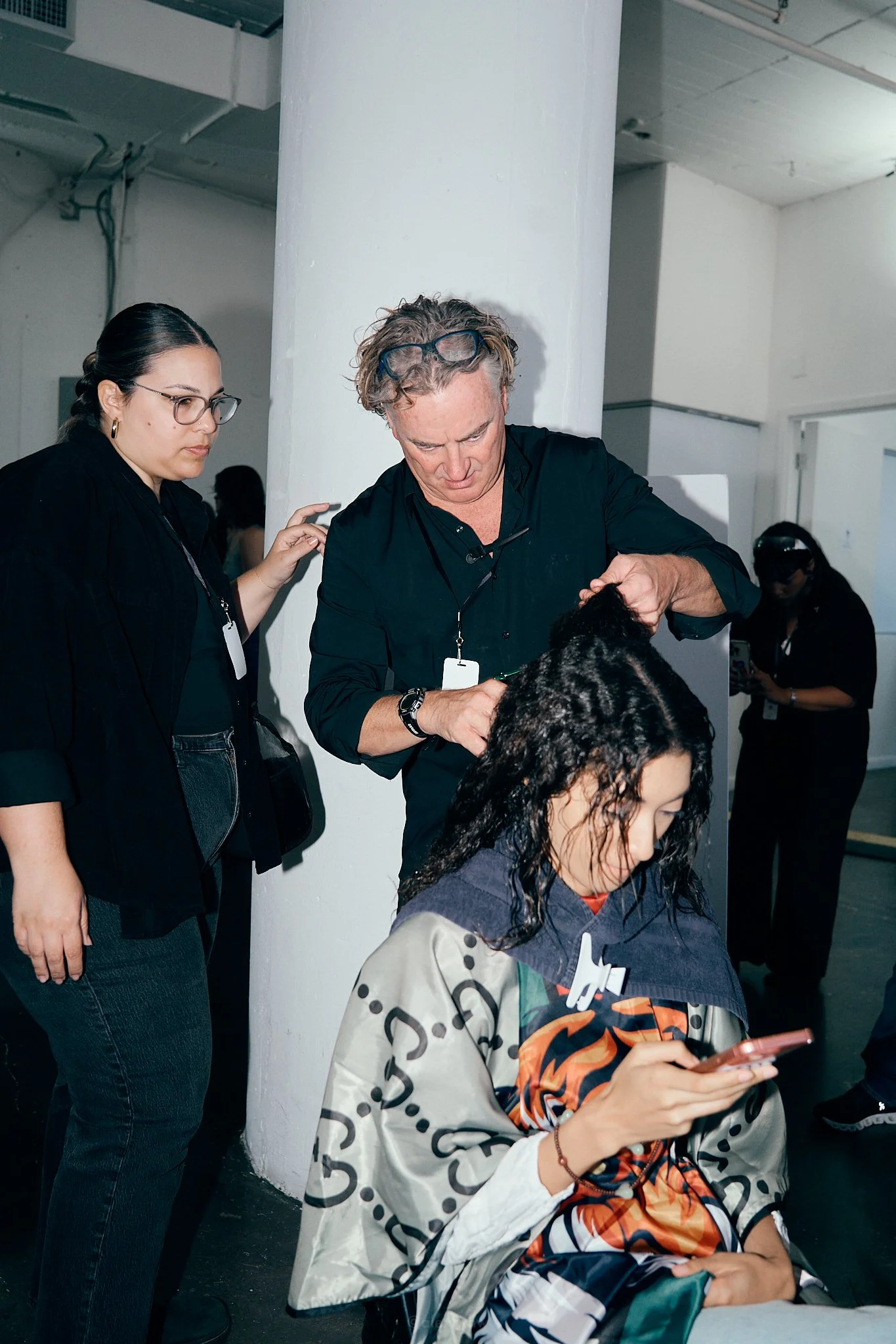 A person getting their hair styled by a hairstylist, while another person looks at their phone.