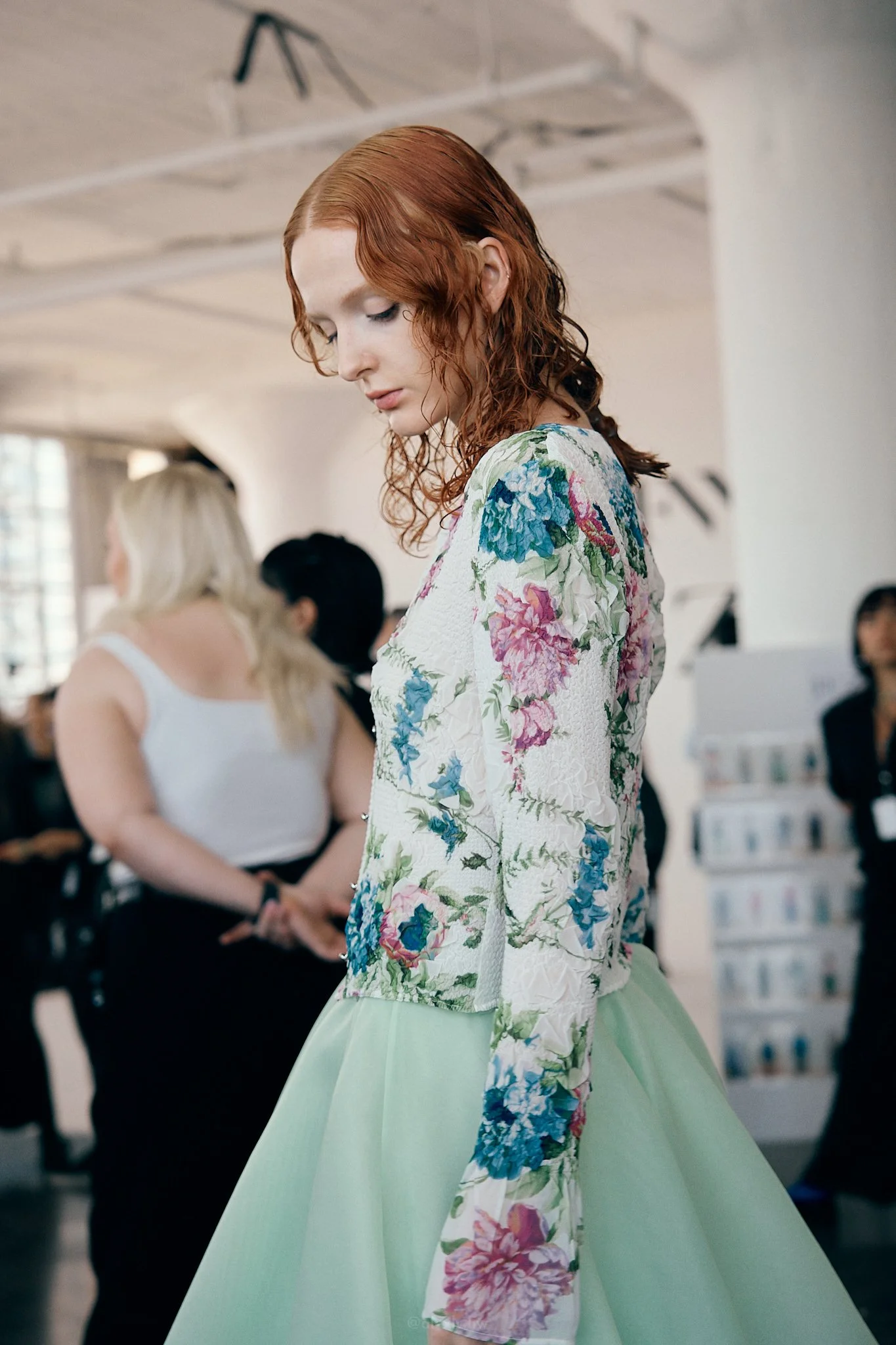 A woman with red wavy hair wearing a floral long-sleeve top and a light green skirt, standing indoors with other people in the background.