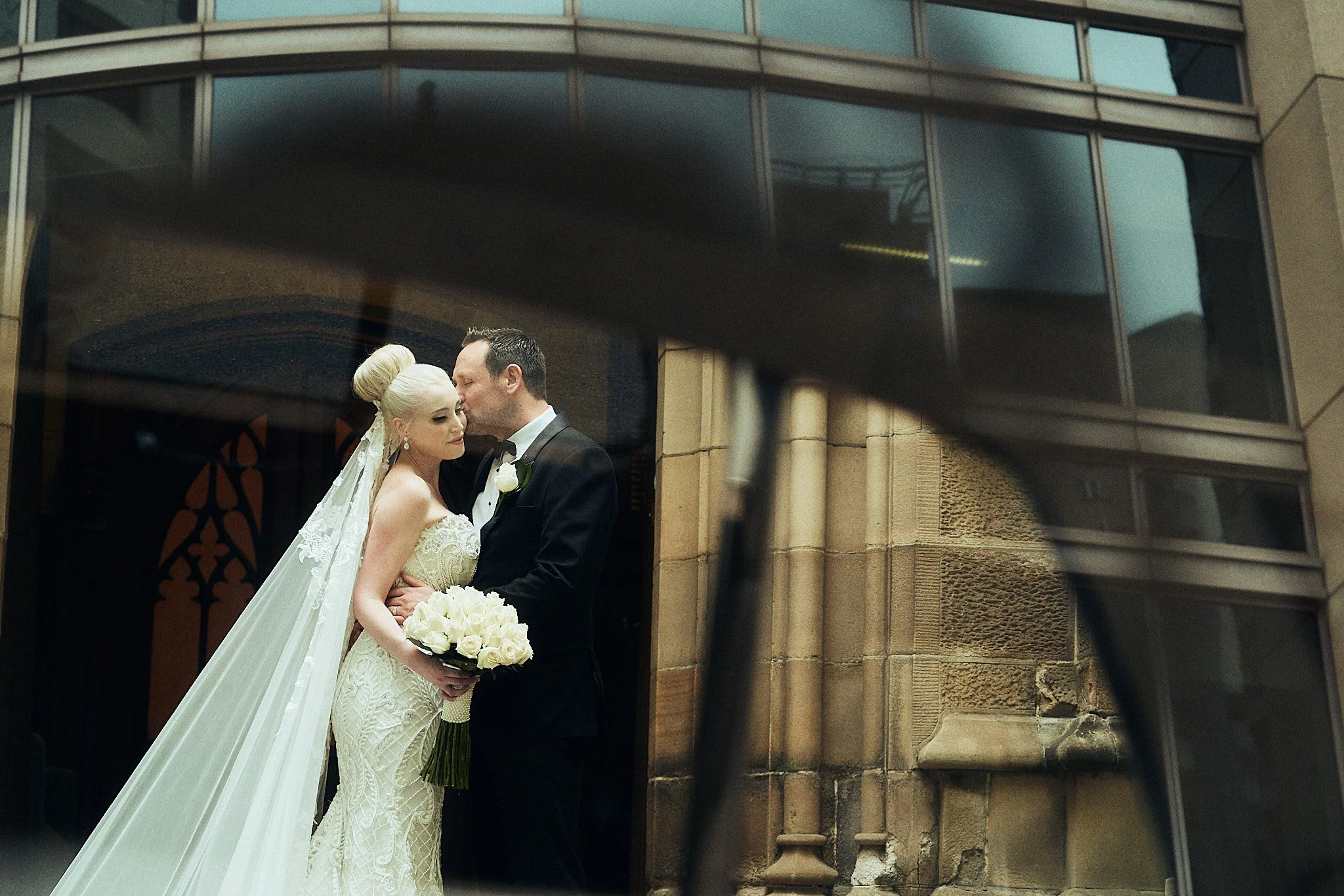 Wedding couple standing close together, with the bride holding a bouquet of white roses, captured through a camera lens filter, outside a modern building.
