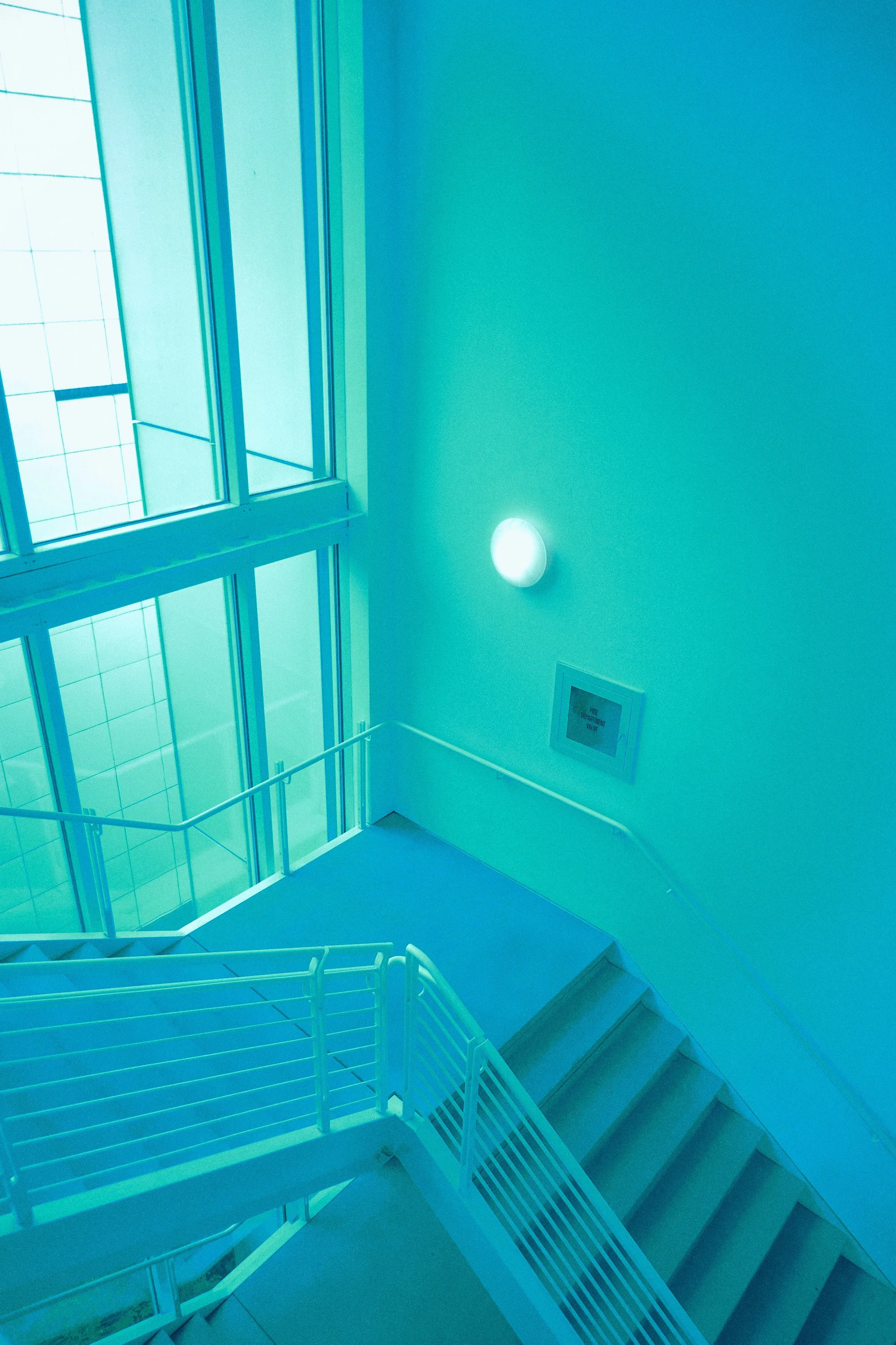 A staircase inside a building with large glass windows, white railings, and a digital sign on the wall, illuminated by blue-tinted lighting.