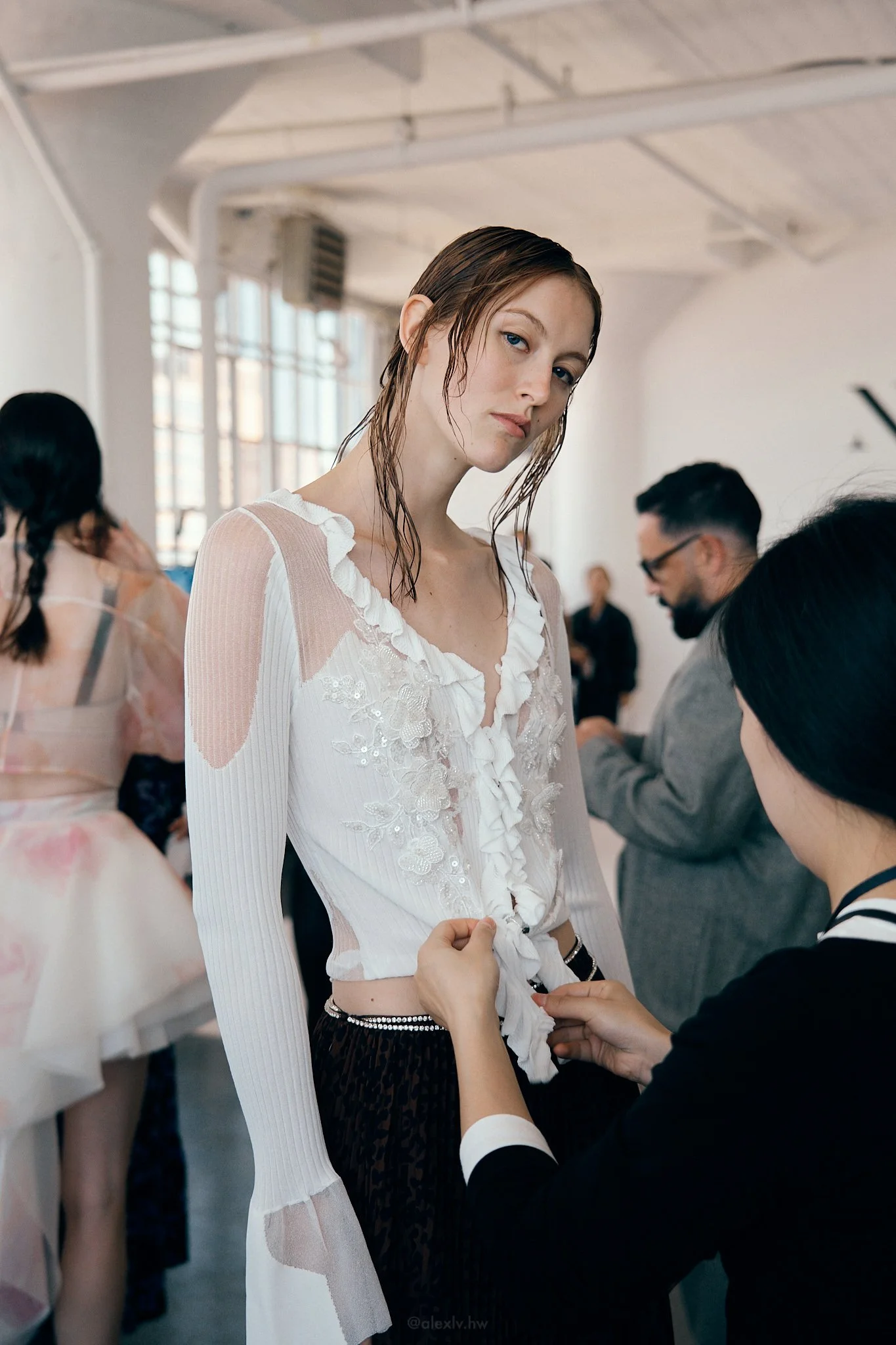 A woman with wet, wavy hair and a white ruffled blouse is having her outfit adjusted by another woman at a fashion event. Other people are visible in the background.