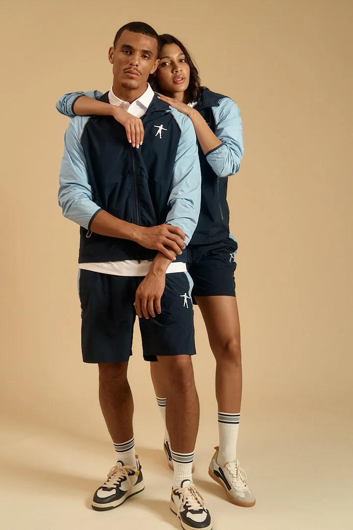 A man and woman dressed in sporty clothing, standing together against a plain beige background. The man is in front, with his arms crossed, wearing a navy blue and light blue windbreaker, navy shorts, striped white socks, and athletic shoes. The woma