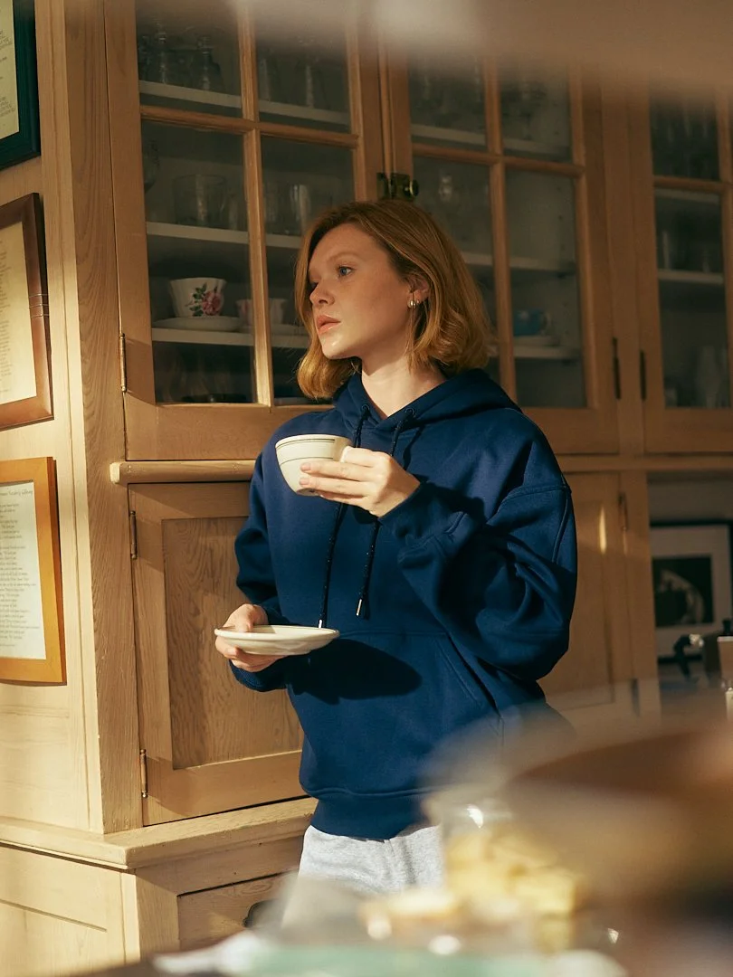 A woman with shoulder-length red hair wearing a navy blue hoodie holds a teacup in one hand and a saucer in the other, standing in a kitchen with wooden cabinets and glass-paneled doors.