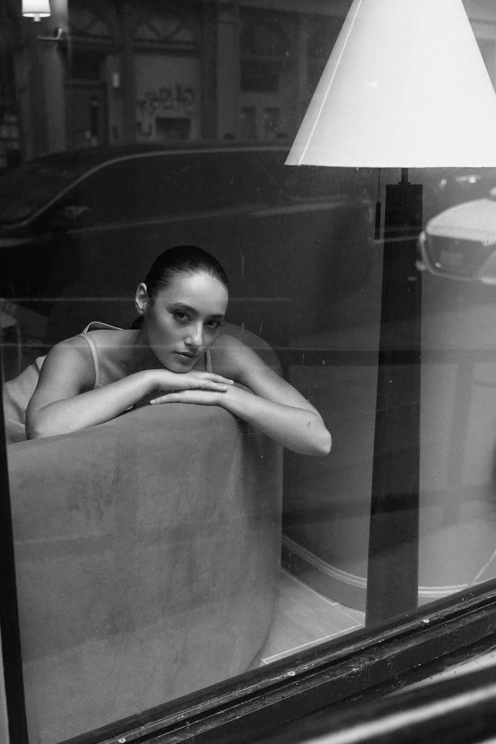 A black and white photo of a woman sitting inside a cafe, resting her head on her crossed arms on a table, looking through a window with reflections of the street outside.