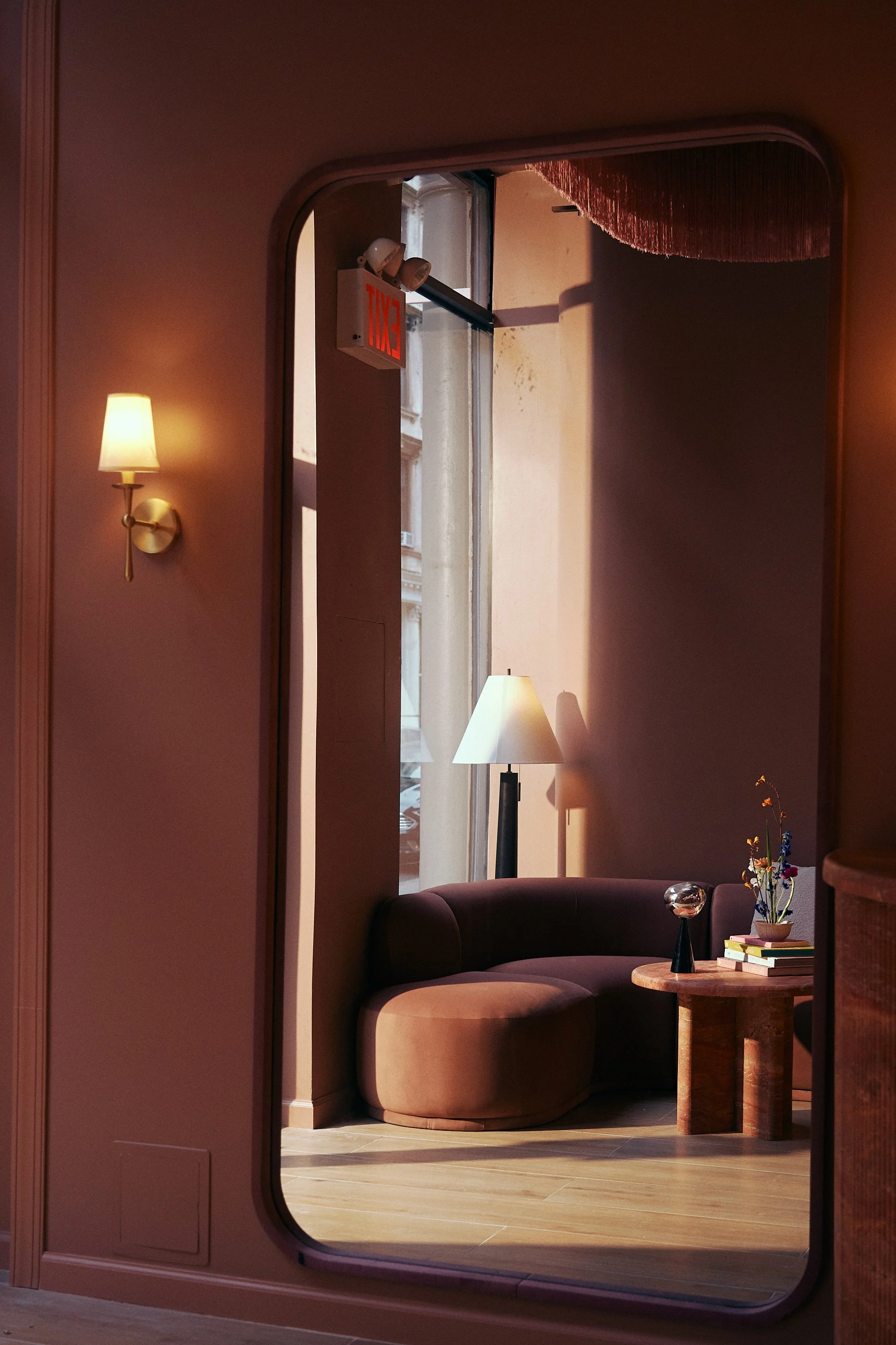 A cozy seating area with a rounded, dark purple sofa, a wooden side table with books and a small vase with flowers, a standing lamp, and a wall-mounted sconce, seen through a doorway with an arched top.