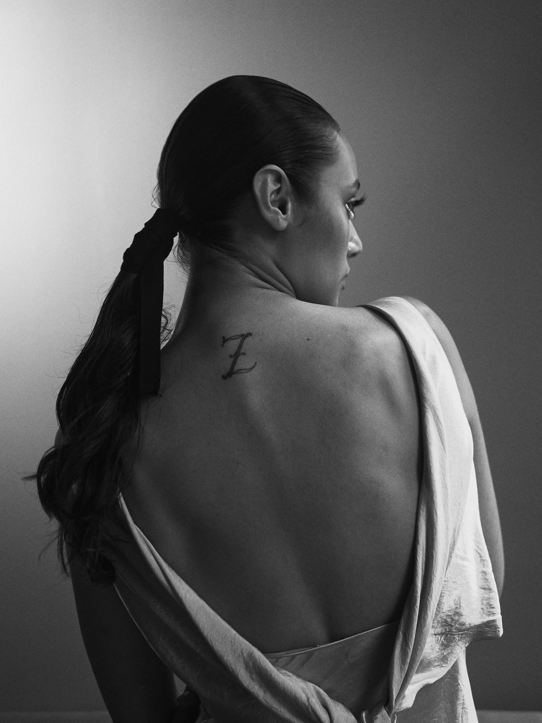 A black and white photo of a woman with long dark hair tied back, looking to her right, showing her back and profile. She has a tattoo of the letter 'Z' on her upper back.