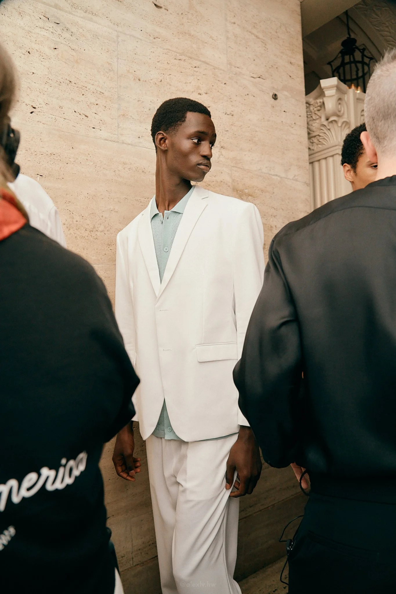 A young man in a white suit standing in a group, looking to the side, against a beige marble wall in an elegant indoor setting.
