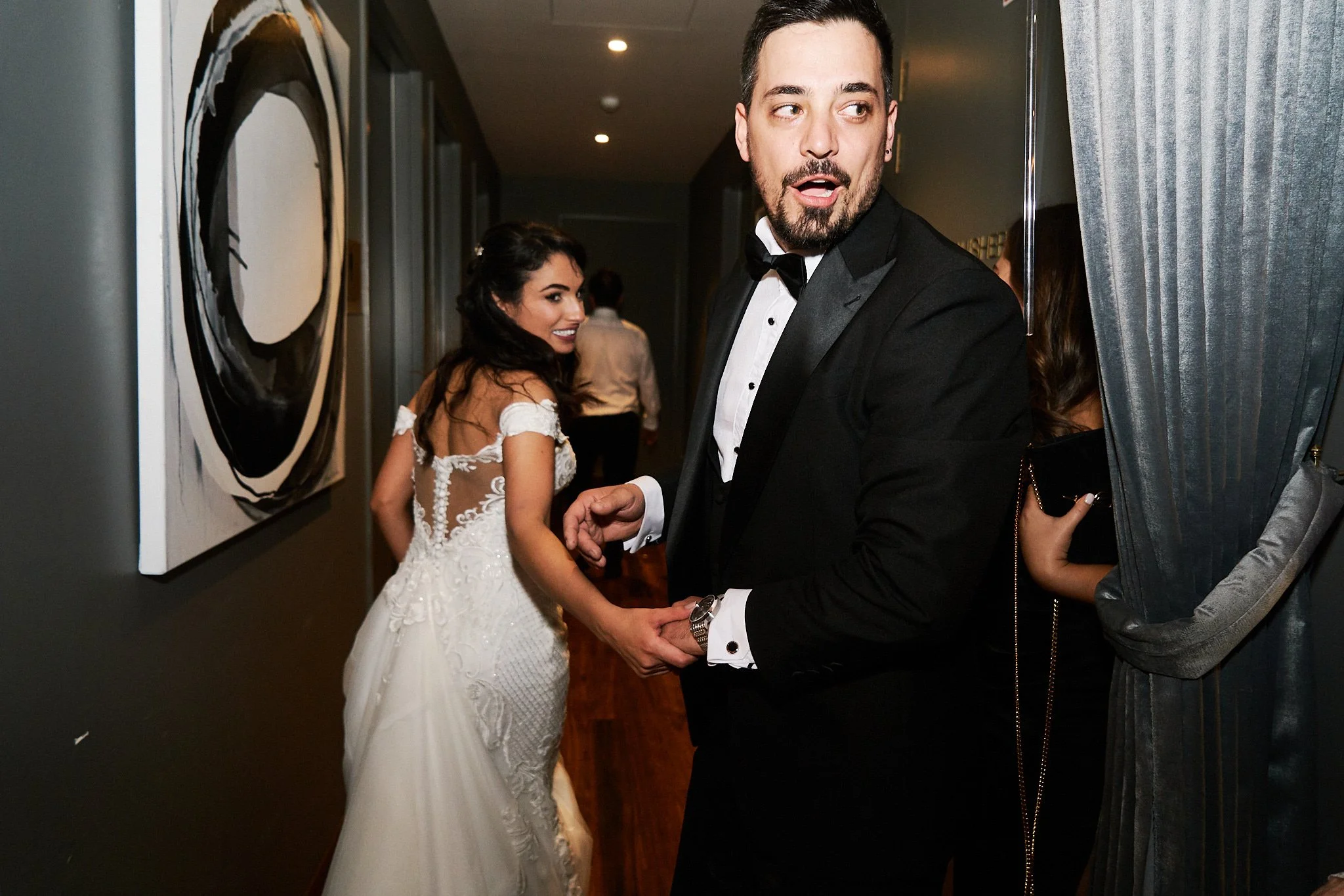 A man in a tuxedo holding hands with a woman in a white wedding dress, standing in an art gallery or exhibition space with dark walls and artwork, and multiple people in the background.