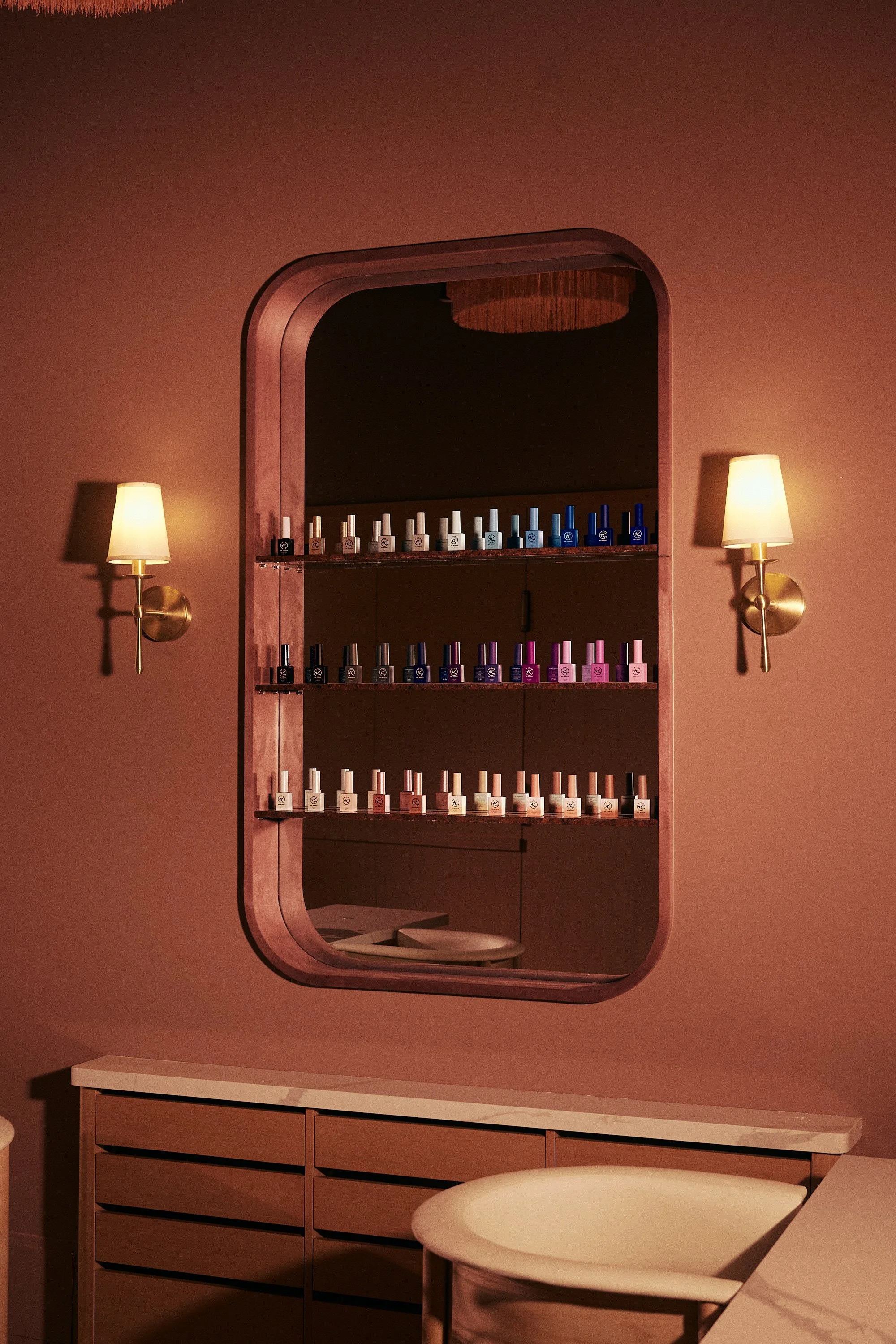 A wall with a mirror displaying nail polish bottles on shelves, and two wall-mounted lamps on either side illuminating the space.