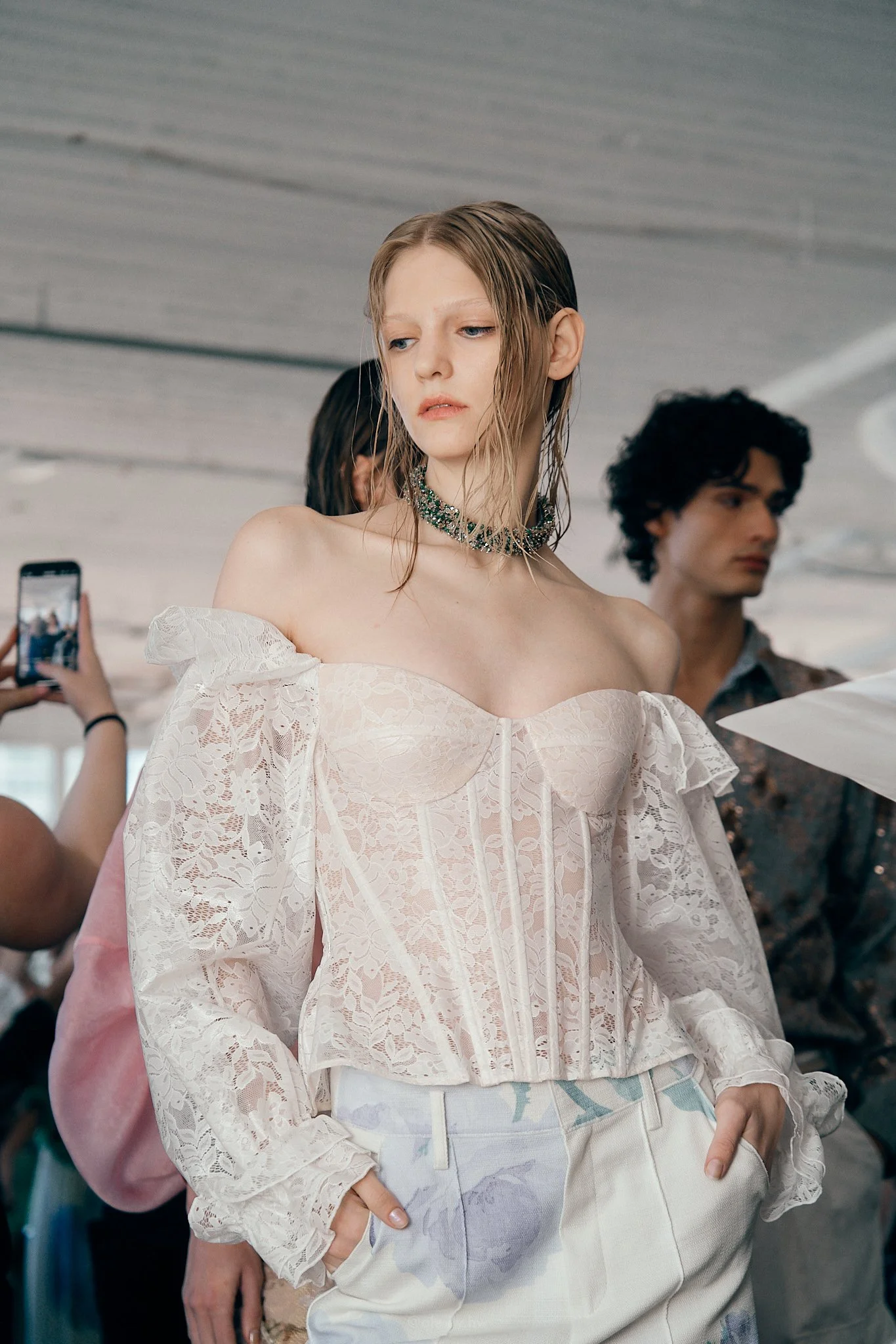 A young woman with wet hair wearing a white off-shoulder lace top and white pants with a floral pattern, standing among others at what appears to be a fashion event or photoshoot.