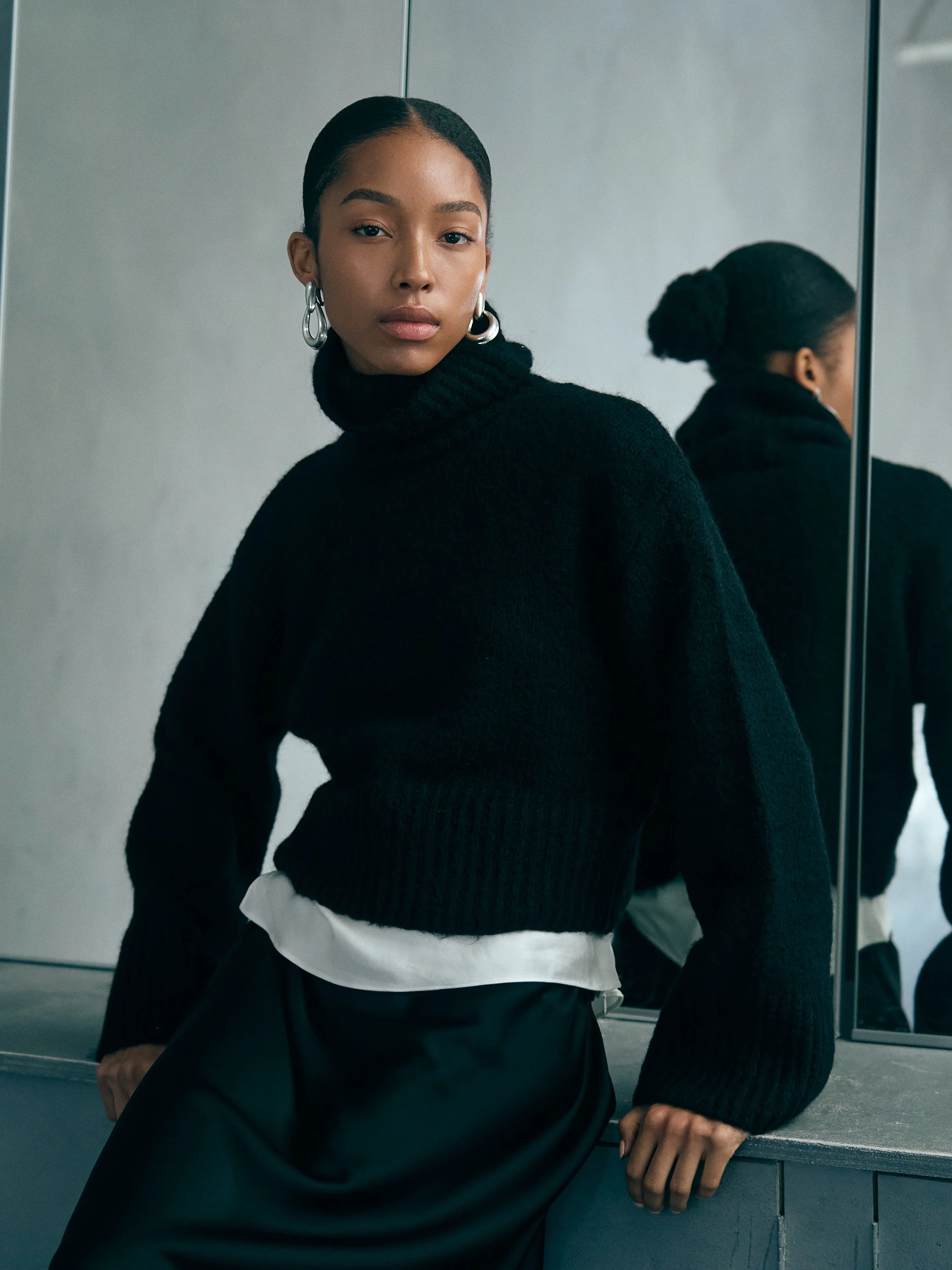 A woman with dark skin and hair styled in a bun, wearing hoop earrings, a black turtleneck sweater, and a white shirt underneath, sitting by a mirror in a modern interior.