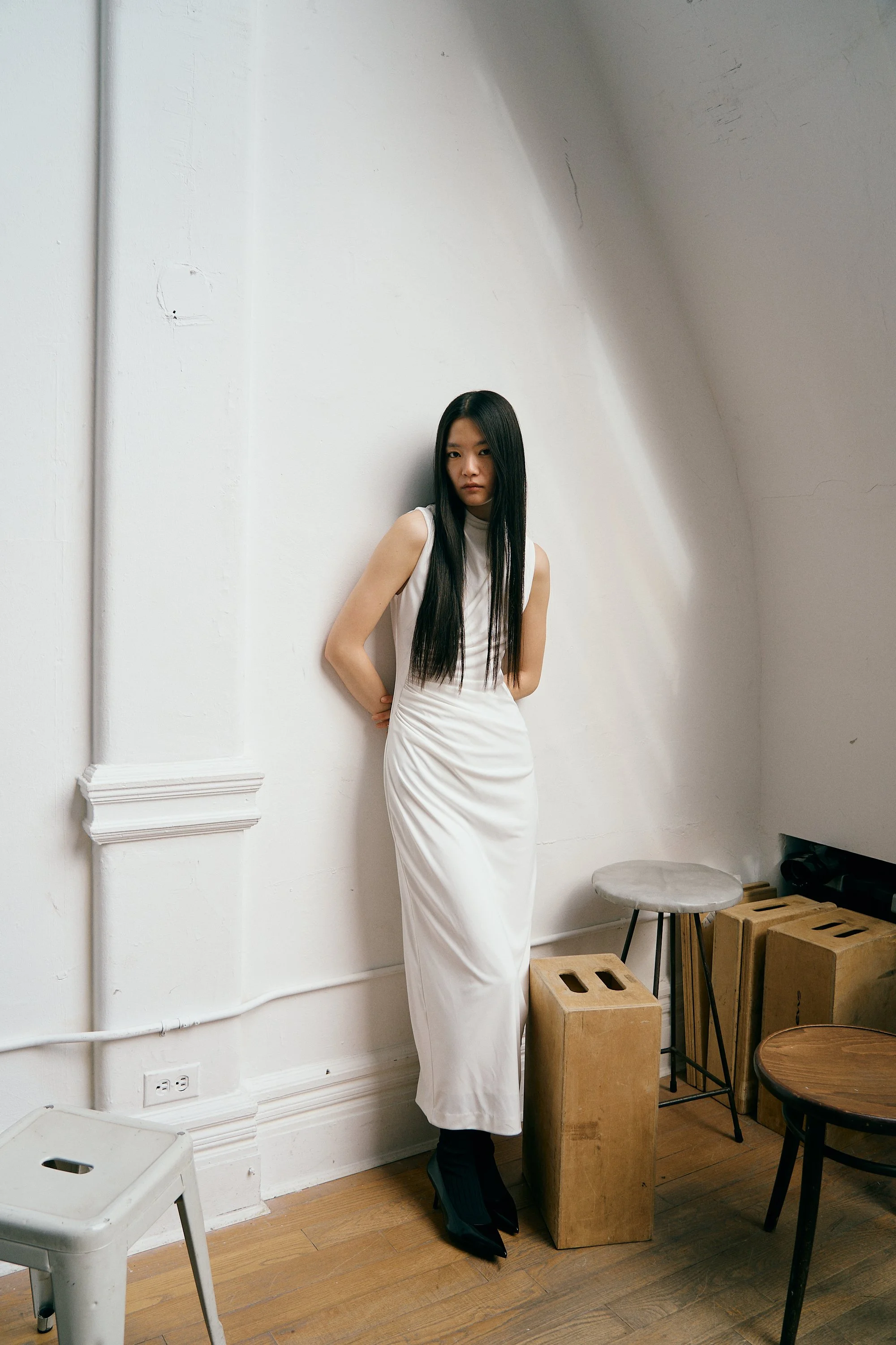 A woman with long black hair wearing a sleeveless white dress and black high heels stands with her back against a white wall in a minimalistic room. The room has wooden floors, a gray stool, and concrete blocks.